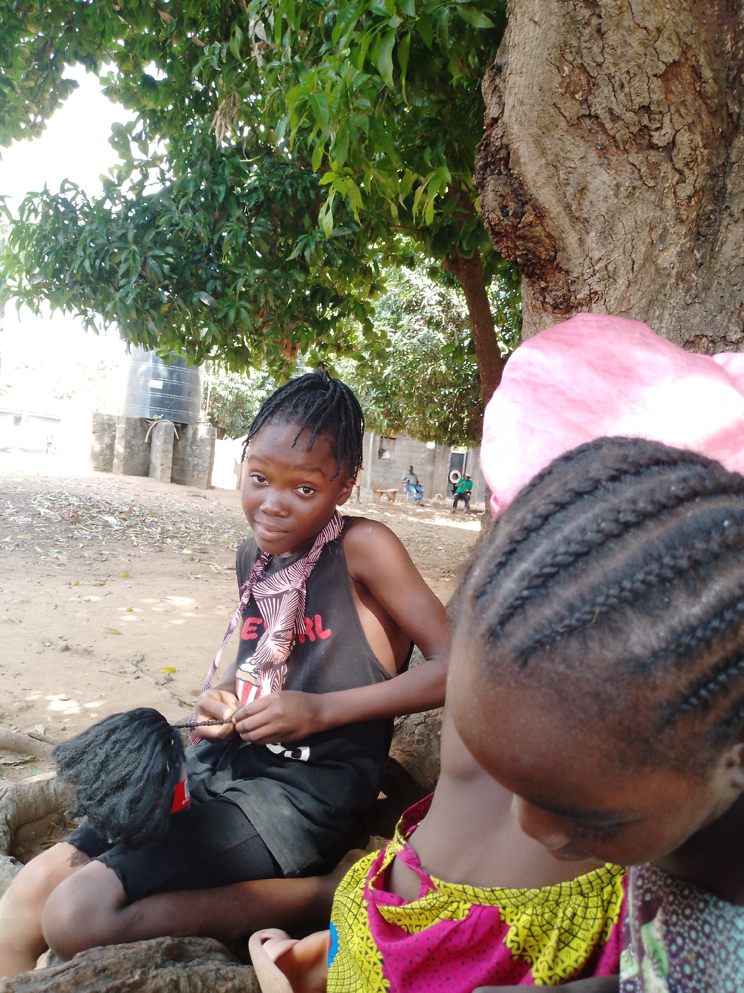 The image captures a scene in an outdoor location. A young girl with braided hair sits in the foreground, wearing a black tank top with a patterned scarf around her neck and black shorts. She is braiding a black wig with a red tag attached. Another child with braided hair is partially visible in the lower right of the frame, wearing a brightly colored yellow and pink dress. Both children are seated near the base of a large tree with thick, textured bark. A dense canopy of green leaves overhead provides shade. In the background, a water tank sits near a simple structure. In the distance, two people are seated in chairs. The ground is covered in dirt and fallen leaves.