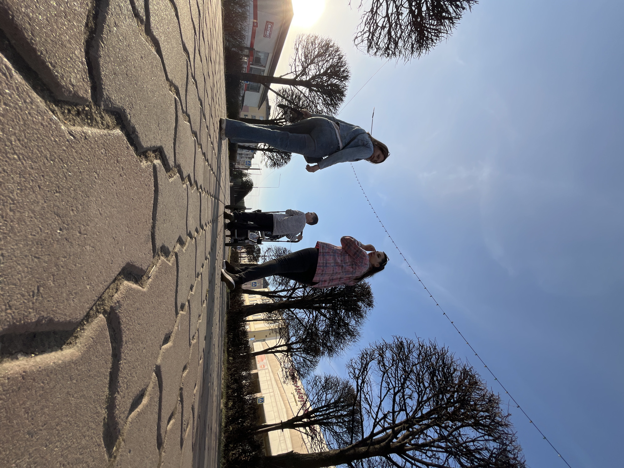 The image depicts a sunny outdoor scene with several people on a sidewalk. A woman in a pink plaid shirt is looking up, while another person in a denim jacket is further away. A third person in a striped shirt appears to be pushing a stroller. Leafless trees line the sidewalk, and buildings are visible in the background. The location is Zelenogorsk, Russia. The ground is paved with interlocking bricks. A string of lights hangs above. The scene conveys a casual, everyday moment.