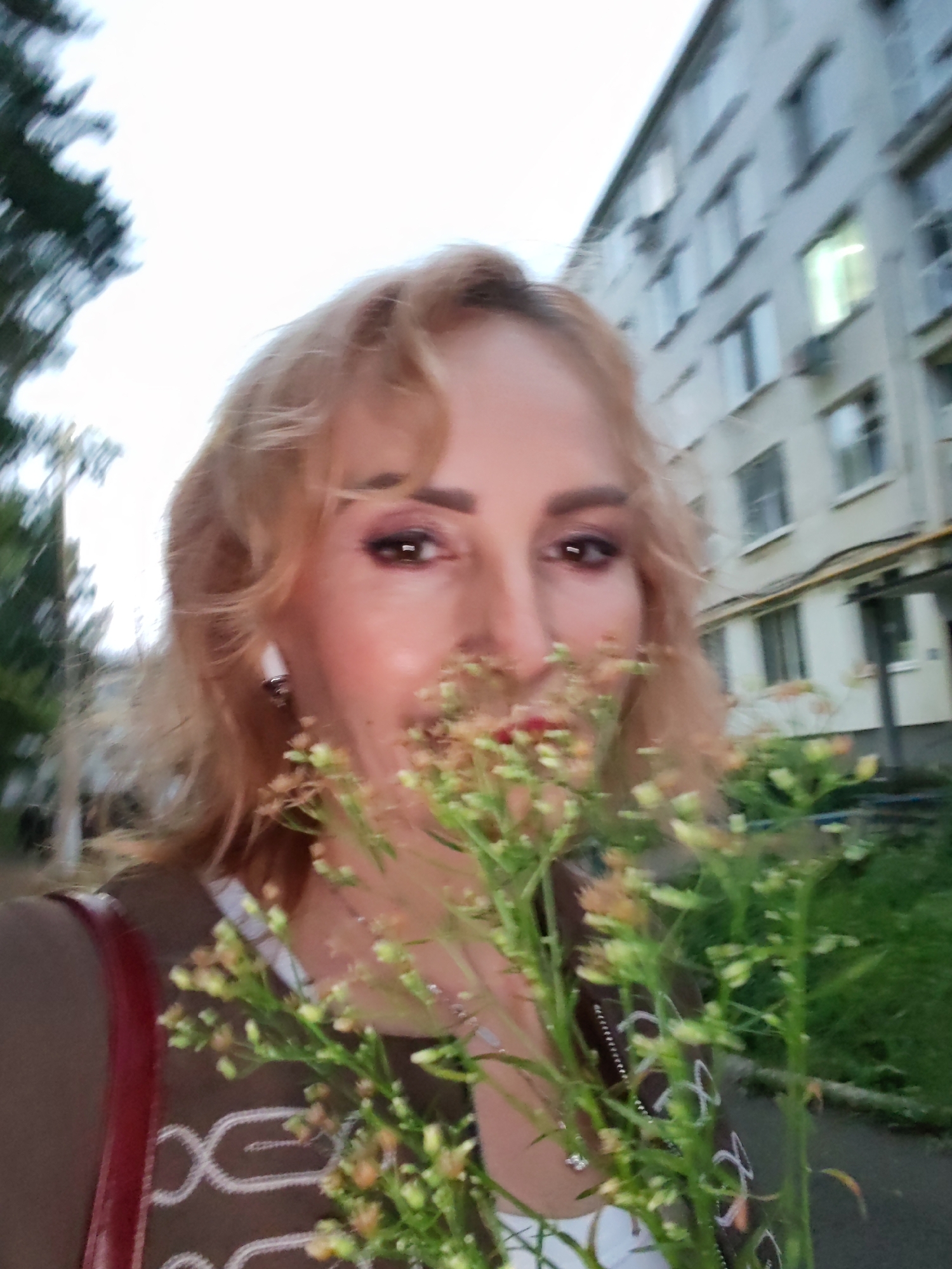 A woman with blonde, wavy hair is holding a small bouquet of wildflowers in front of her face. She is wearing a brown top with a white shirt underneath, and a red strap from a purse is visible on her left shoulder. She has on eye makeup and red lipstick. The background shows a multi-story apartment building and some greenery. The image appears to have been taken in Chaikovsky, Russia, as indicated by the characteristic architecture of the apartment building and the overall setting. The lighting suggests it might be late afternoon or early evening.