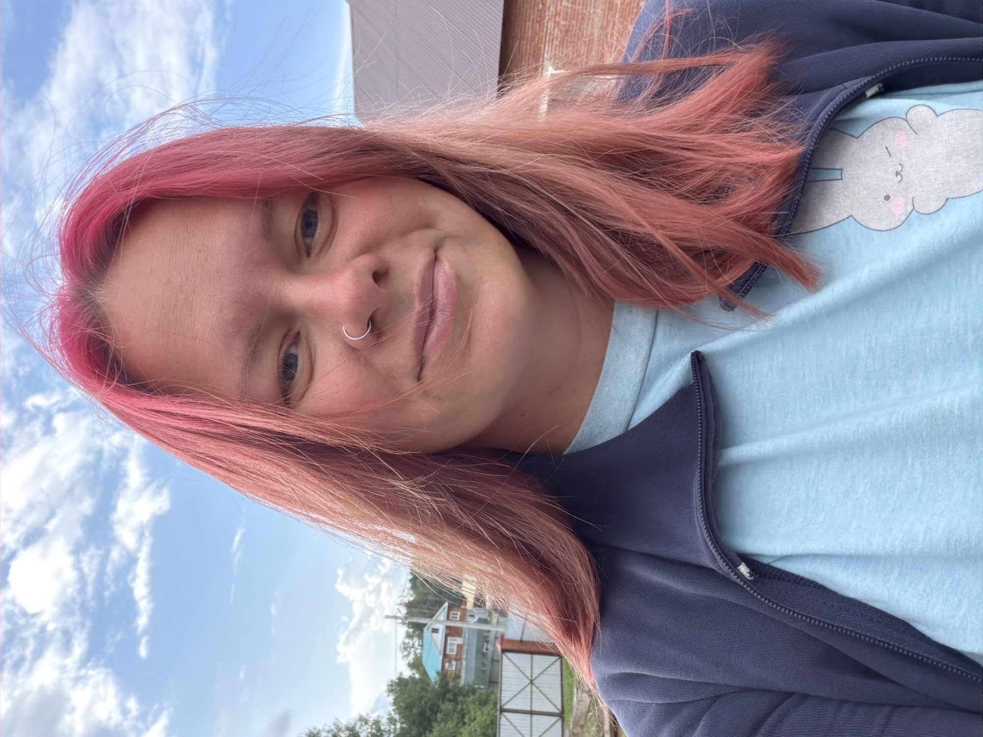 A young woman with bright pink hair and a nose ring is taking a selfie outdoors. She is wearing a light blue t-shirt with a cartoon bunny graphic and a dark blue zip-up jacket. In the background, there is a blue sky with scattered clouds, trees, a fence, and a brick building. The setting appears to be a residential area, possibly in the city of Chaikovsky, Russia, given the architectural style of the houses and the general environment.