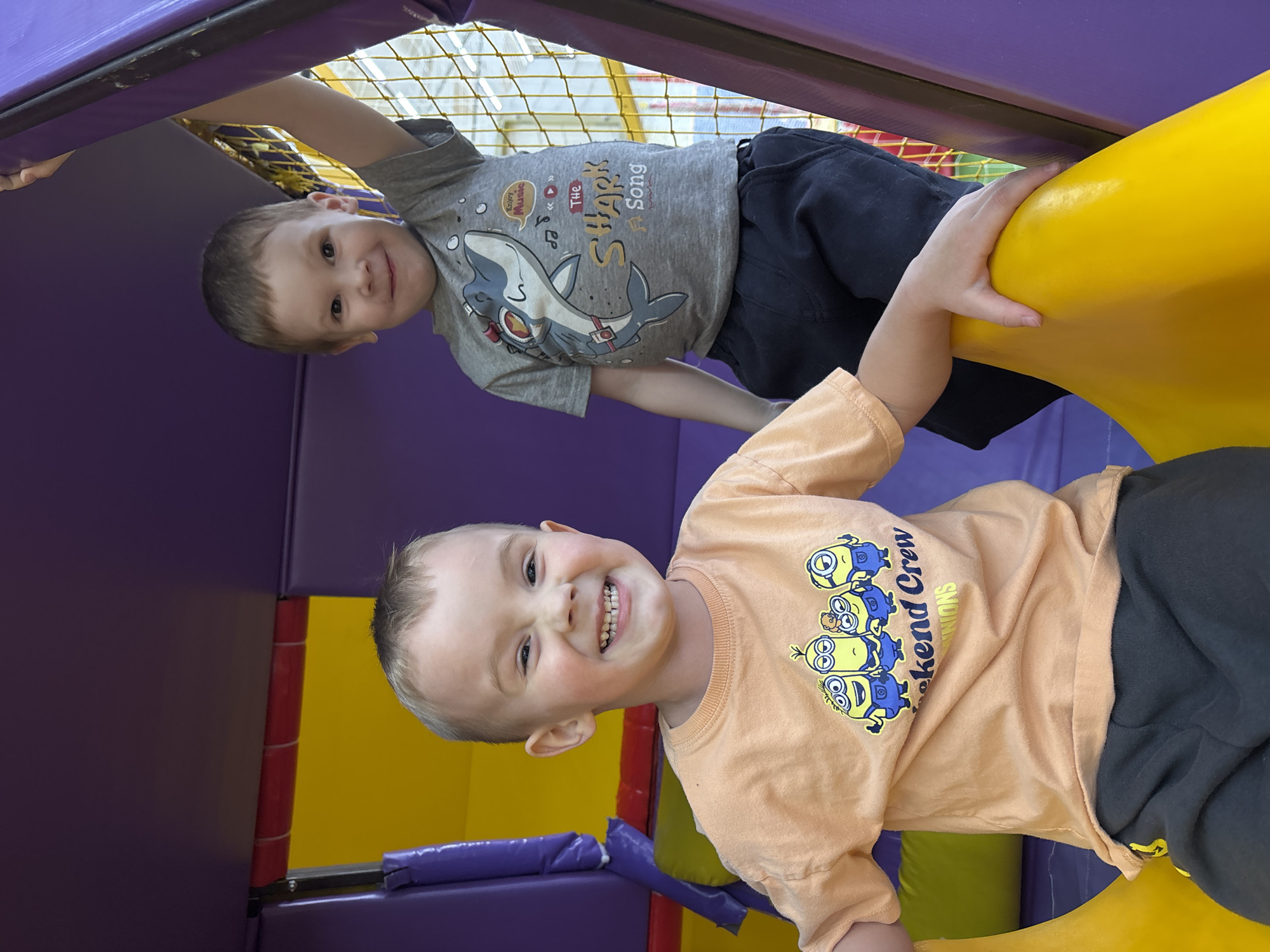 Two young boys are playing in an indoor playground. One boy is wearing an orange t-shirt with Minion graphics and dark-colored pants. The other boy wears a gray t-shirt with a shark graphic and dark-colored pants. They are both smiling and appear to be having fun. The playground equipment is colorful with purple, yellow, and green elements. The location in the city of Chaikovsky, Russia is not discernible from the image.