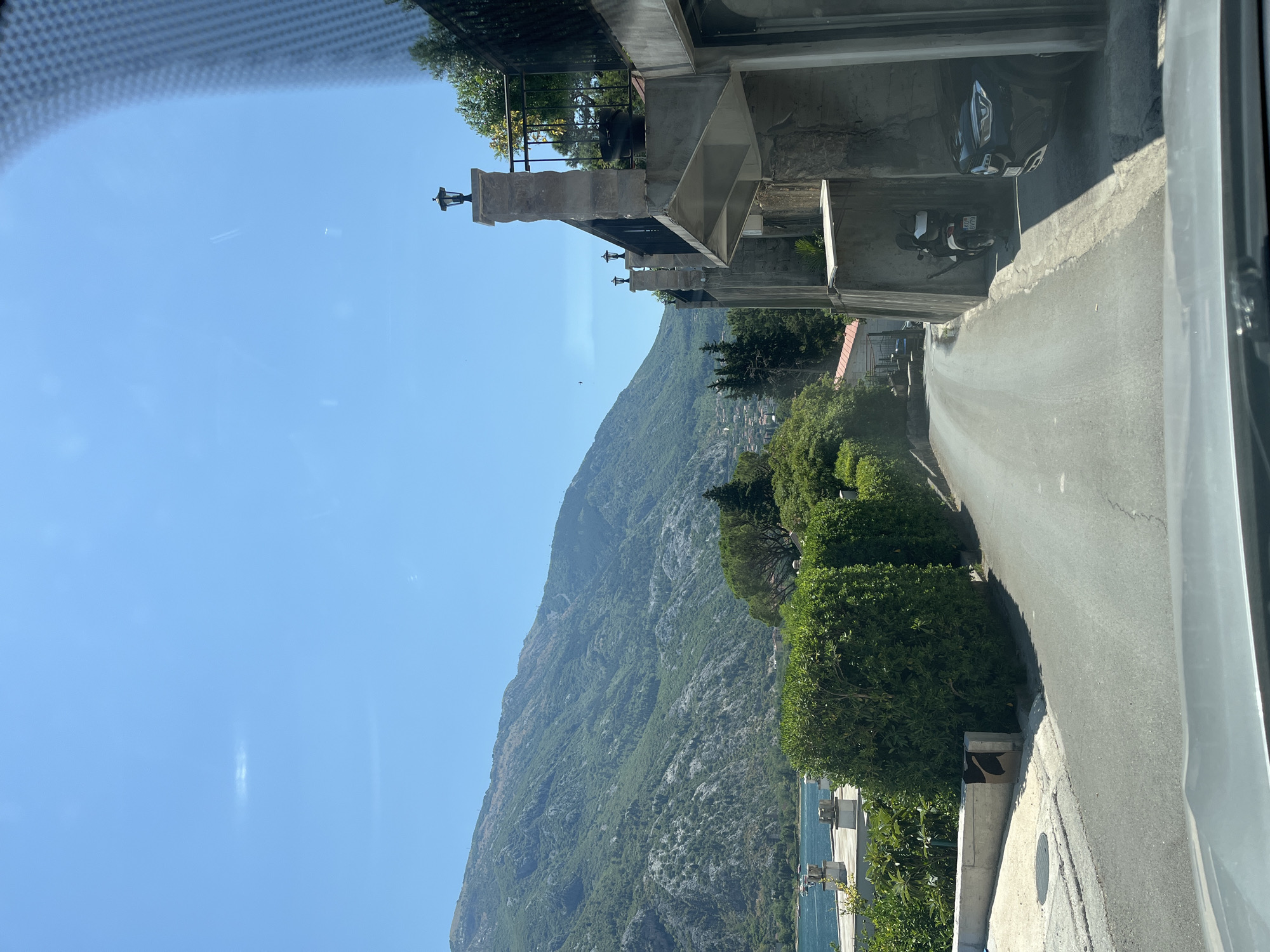 A clear, sunny day in Muo, Montenegro. The image captures a winding asphalt road leading uphill. On the right, a multi-story stone and concrete structure is visible, featuring balconies with ornate lampposts and railing. A black Mercedes-Benz SUV is parked to the right of the road, partially obscured by the structure. A white scooter is parked in the alcove of the building. The road is lined with neatly trimmed green hedges on the left. In the distance, a body of water, likely the Bay of Kotor, is visible, with a mountainous, tree-covered landscape rising behind it. The sky is a vibrant, cloudless blue, with faint white streaks that may be contrails from aircraft. The image appears to be taken from inside a vehicle, with the upper portion of a windshield and its defroster lines visible in the foreground.