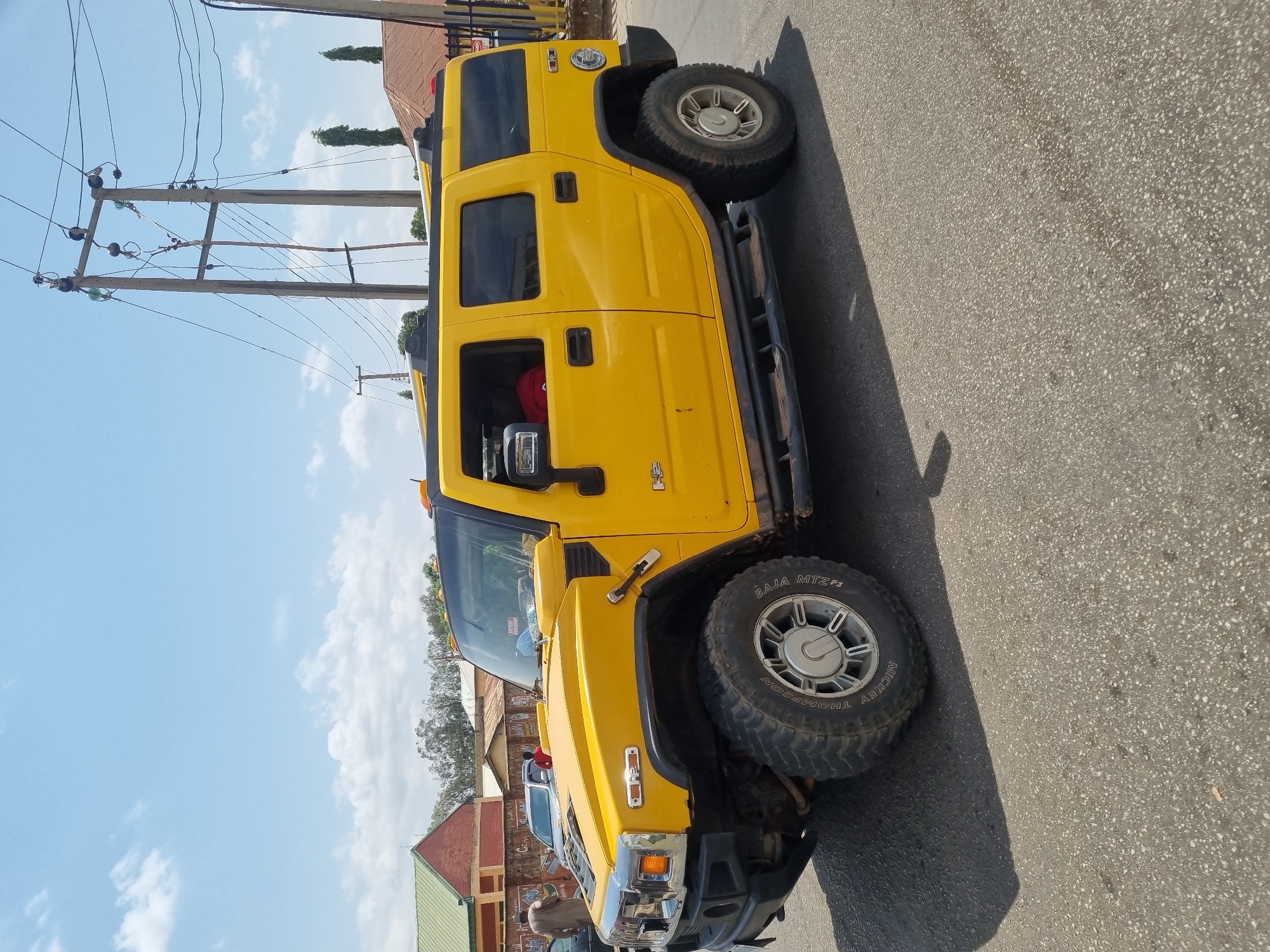 A yellow Hummer H2 is parked on a paved road in Jos, Nigeria. Overhead power lines are visible above the Hummer. Houses and trees are in the background. The sky is partly cloudy.