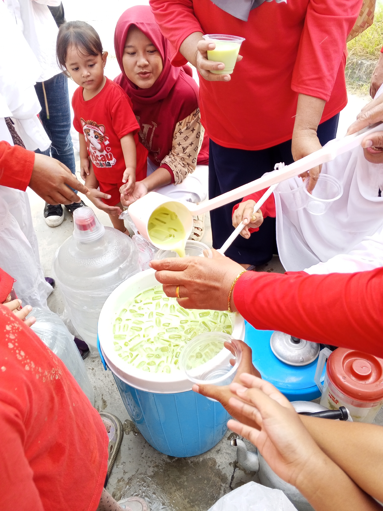 An outdoor scene in Ciruas, Indonesia, features a group of individuals, predominantly wearing red, gathered around a large blue cooler and a large water jug. A young child in a red shirt is held by a woman wearing a red hijab. Multiple people are holding cups, and some are pouring a light green beverage from a pitcher into the cooler, which is filled with ice and the same green beverage. Another person holds a translucent cup with the green drink. A red-lidded container is visible to the right. The ground is wet, suggesting a recent spill or damp conditions. The overall atmosphere appears to be a communal event or food distribution.