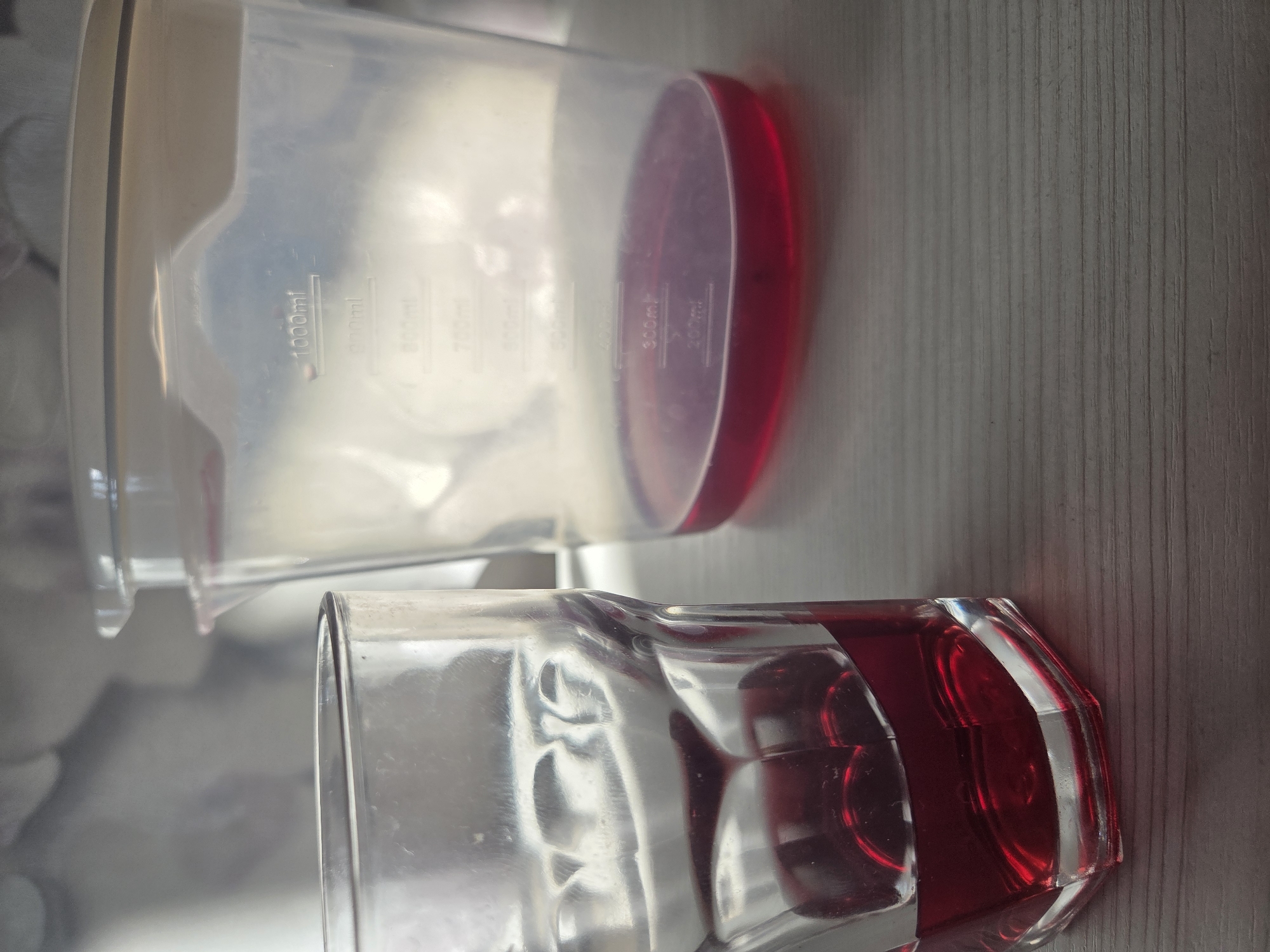 A close-up, eye-level shot depicts a clear measuring cup lying on its side and partially filled with a red liquid. The markings indicating measurements are visible on the side of the cup. Next to it, also lying on its side, is a clear, multifaceted glass partially filled with the same red liquid. Both objects rest on a light-colored, vertically striped surface, possibly a tabletop. No people are present in the scene. The location in the city of Yug, Russia, is not determinable from the image.