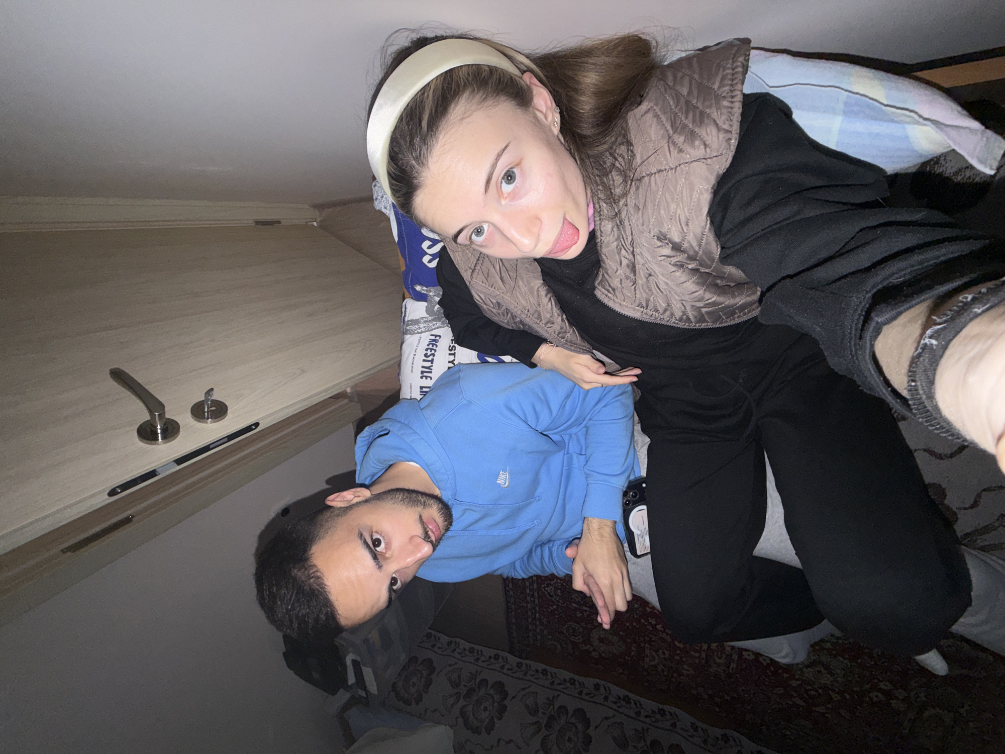 Two individuals are present in the image. A woman with a headband is wearing a brown vest and black clothing. She is sticking out her tongue and her hair is brown. A man is wearing a blue sweatshirt. The background includes bedding and a patterned rug. A door and ceiling are visible. The location is potentially a residential interior based on the objects present, and can not be definitively identified as being located in Milevo, Bulgaria.