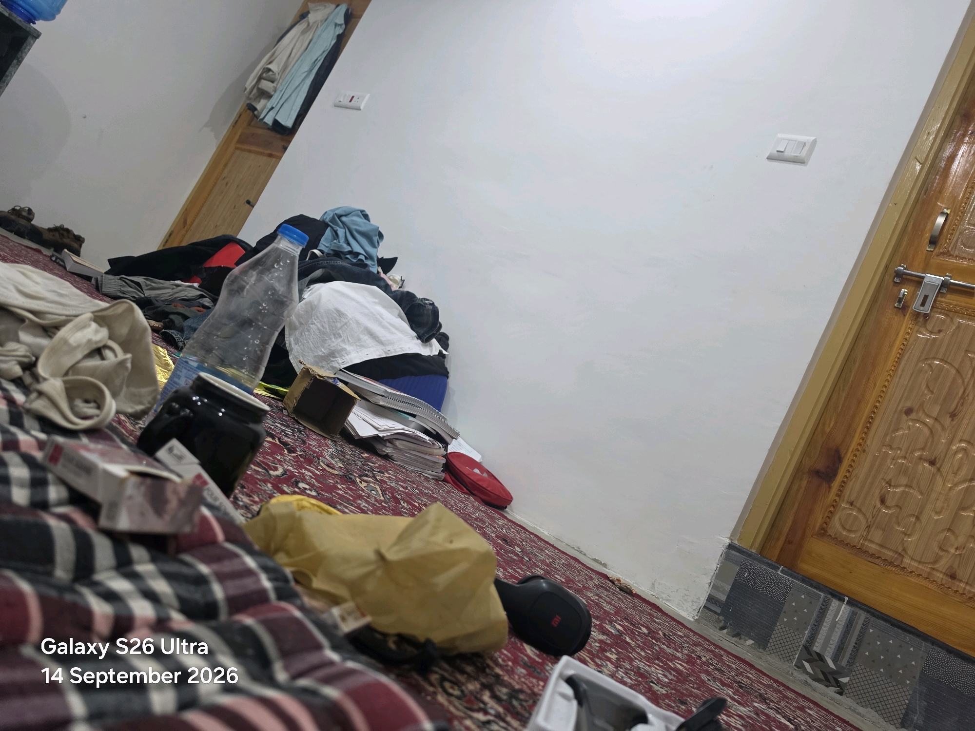 The image depicts a cluttered room, likely a bedroom or living space, with items strewn across the floor and surfaces. The floor is covered by a red patterned rug. In the foreground, on the rug, is a partially visible white and red plaid blanket or fabric. To the left of this, a white and red cigarette pack is visible, along with other personal items. Further back, a clear plastic water bottle with a blue cap rests near a black ceramic mug. A stack of books and papers is piled near a red bag. Clothing, including a blue t-shirt, is draped over various objects and surfaces. A wooden door with decorative carvings is visible on the right side of the image, with a metal door handle and lock. Electrical outlets are present on the white wall. The overall impression is one of disarray. The image is dated "14 September 2026" and tagged "Galaxy S26 Ultra." There are no people visible in the image. The location is identified as Kulgam, India.