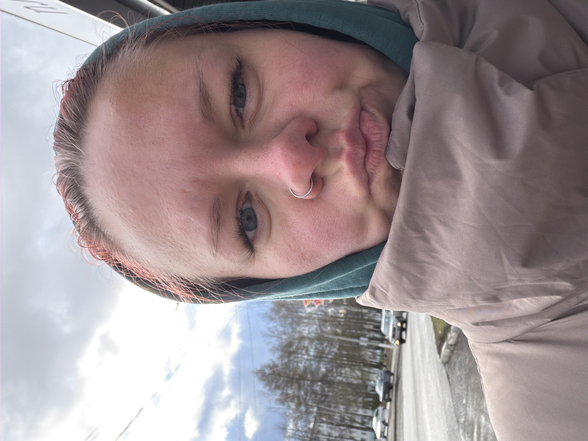 A close-up selfie shows a person's face in a playful pose, making a "duck face" with pursed lips, and looking directly at the camera. The image is rotated 90 degrees counter-clockwise, presenting the subject's head horizontally. The person has light skin and eyes, and wears a silver nose ring in their left nostril. Light-colored hair, possibly with subtle reddish or pinkish streaks, is visible around the forehead, tucked under a teal or green hooded garment. They are also wearing a light brown or beige puffer jacket.
The background reveals an outdoor setting, suggesting the selfie was taken on a street in Chaikovsky, Russia. A road with several cars and bare trees lines the side of the street, indicating a season like late autumn, winter, or early spring. The sky is overcast and bright, consistent with daytime. No discernible text or specific landmarks of Chaikovsky are clearly visible in the background, which is somewhat blurred. The overall impression is casual and lighthearted.