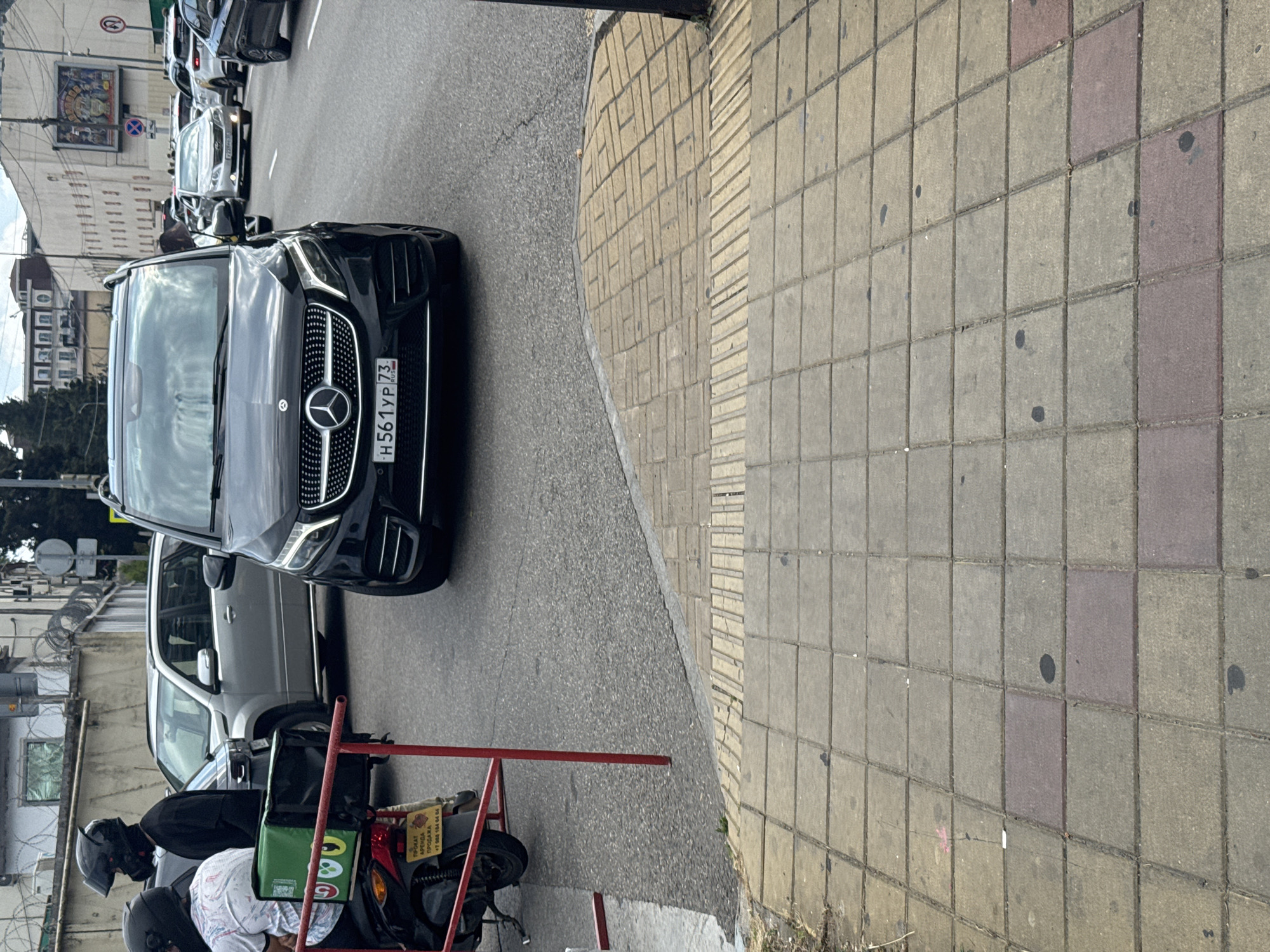 The image shows a street scene in Sochi, Russia, with several vehicles and a sidewalk in the foreground. A black Mercedes-Benz V-Class van is prominently positioned in the center of the frame, facing forward and displaying the license plate "H561YP 73". To the left of the van, a silver SUV is visible, and further back, other cars are in traffic. A motorcycle with a green delivery box and two individuals wearing helmets are parked on the left side. The sidewalk in the foreground is paved with concrete tiles and has tactile paving elements. The environment appears to be urban, with buildings visible in the background and overhead wires.