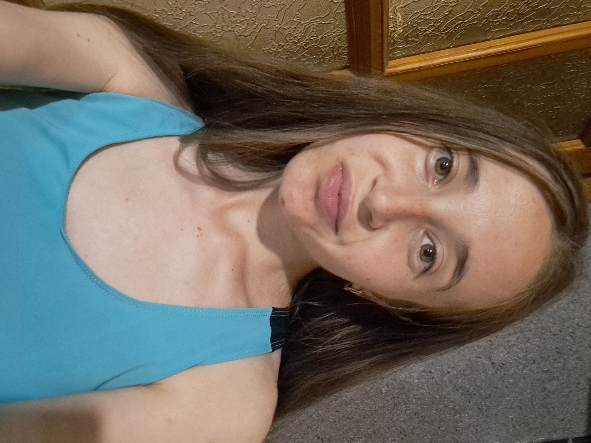 A young woman with long, brown hair is pictured in a close-up selfie. She is wearing a turquoise tank top. Her expression is slightly puckered. The background is blurred, with a textured surface on the left and a darker, rougher surface on the right. The scene appears to be indoors. The location is Vypasne, Ukraine.