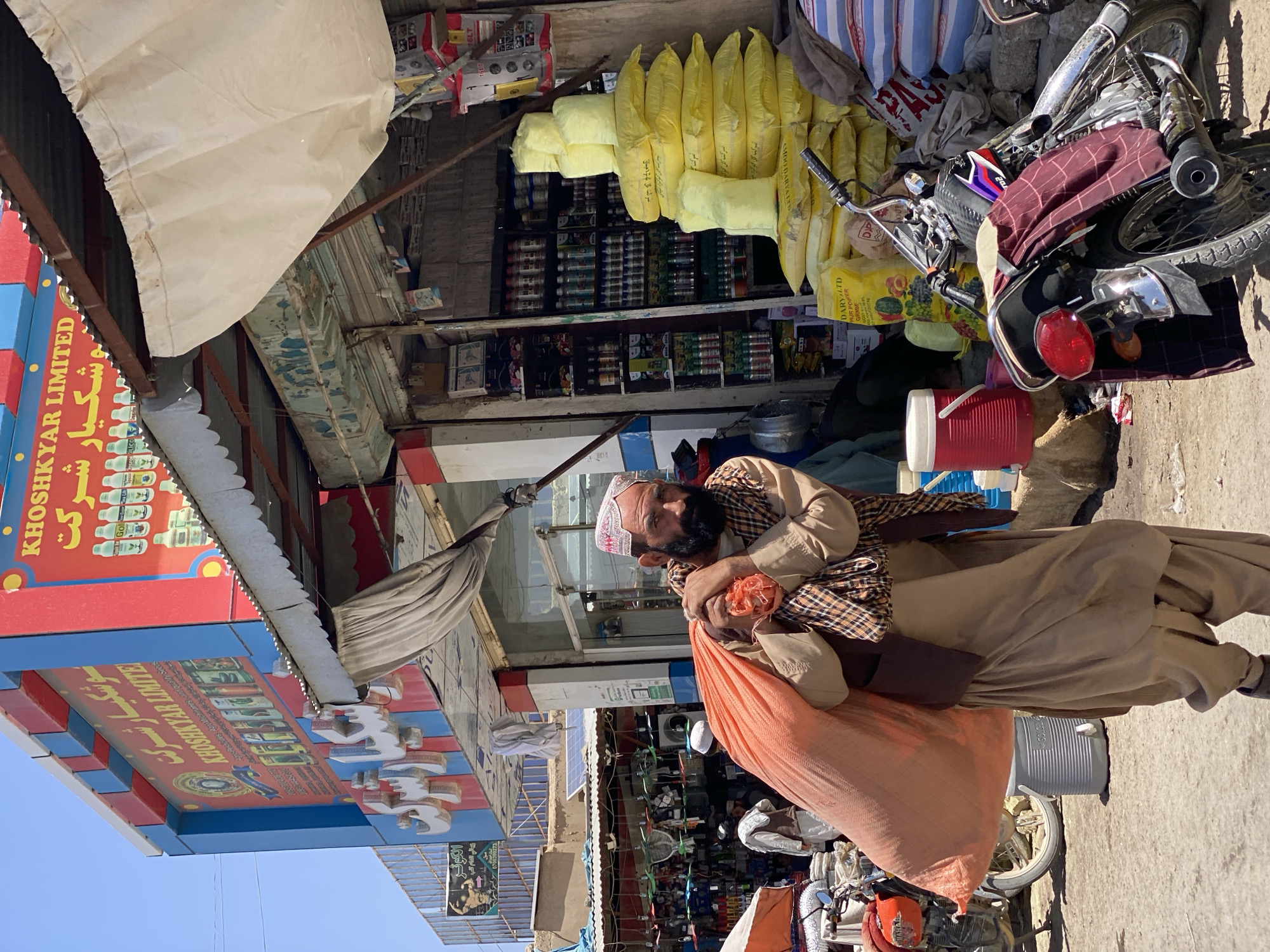 A man, identified as a Pashtun merchant in Kandahar, Afghanistan, walks while carrying a large orange sack of goods over his shoulder. He wears a white skullcap with red trim and a plaid scarf over a tan tunic and trousers. Behind him, a store displays various items, including bags of what appear to be fertilizer or grain, and shelves stocked with bottled products. A motorcycle is parked in the foreground. The scene is a bustling street market, characteristic of Kandahar's commercial areas.