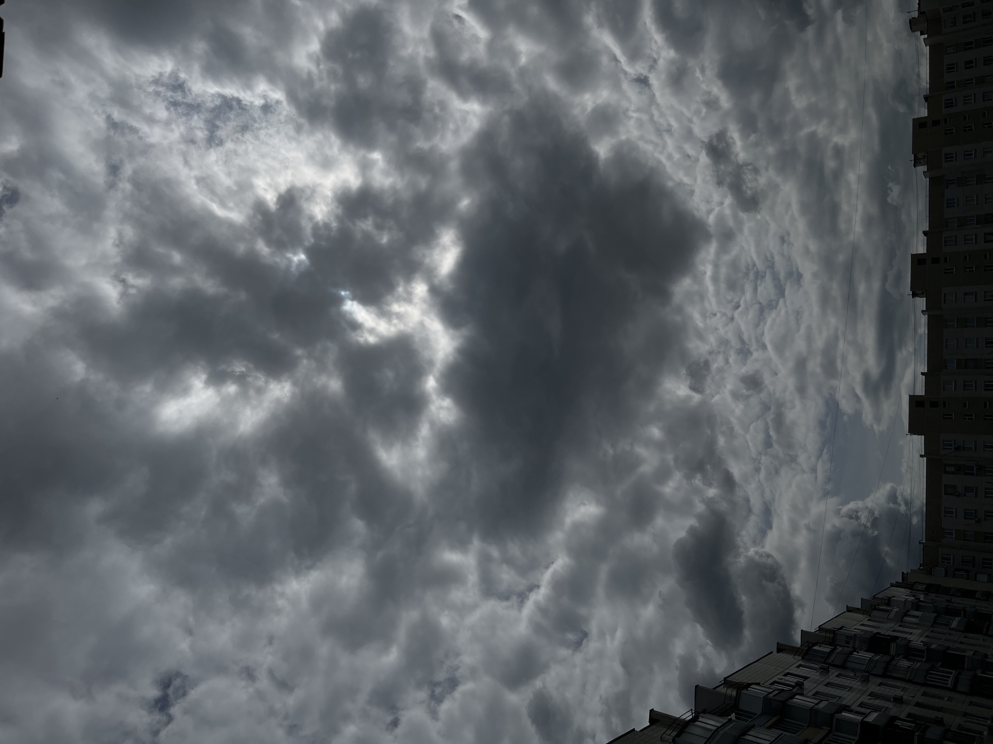 A view of a cloudy sky above apartment buildings, suggesting an overcast day. The sky is filled with dense, dark gray clouds, with patches of brighter light peeking through in some areas, indicating a break in the clouds or the sun trying to shine. Below the sky are the tops of several apartment buildings, typical of urban architecture. The buildings are multi-story, with visible windows and balconies. The scene appears to be in a residential area of a city, likely Moscow, Russia, given the architectural style of the buildings. There are no people or specific actions depicted, only the environment.