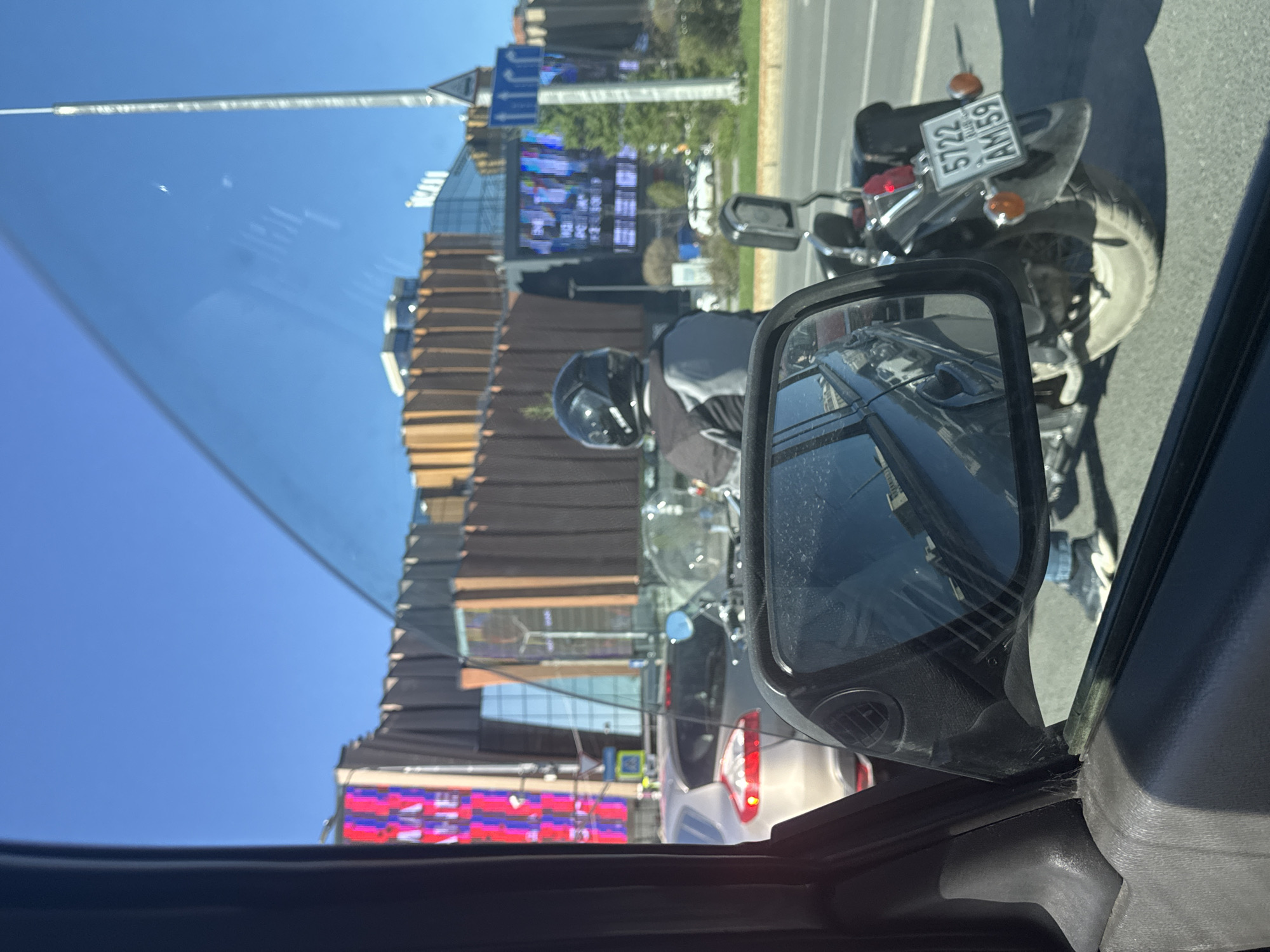 The image, taken from inside a vehicle, shows a street scene in Perm, Russia. The vehicle's side mirror reflects other cars. Outside, a motorcyclist with a black helmet is stopped in traffic, identifiable by the license plate "5722 AM 59 RUS". A large building is in the background, featuring a large electronic screen. The sky is clear and blue.