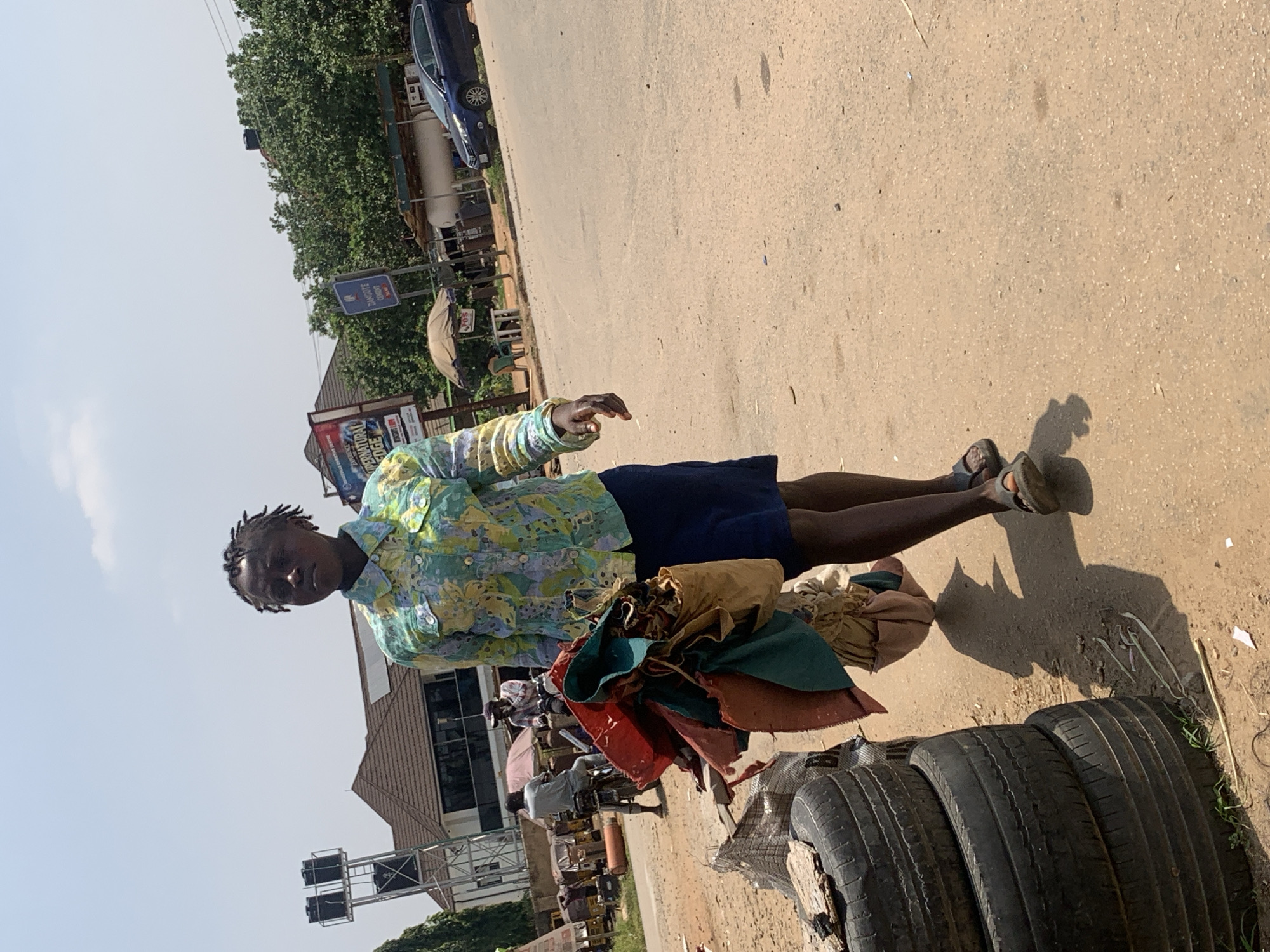 A woman walks across a dusty road in Abuja, Nigeria, carrying a bundle of colorful fabrics. She is wearing a floral print shirt and a dark blue skirt. In the background, there are buildings, trees, and a blue car. Stacked tires are visible on the left side of the image. The sky is clear and bright, indicating a sunny day.