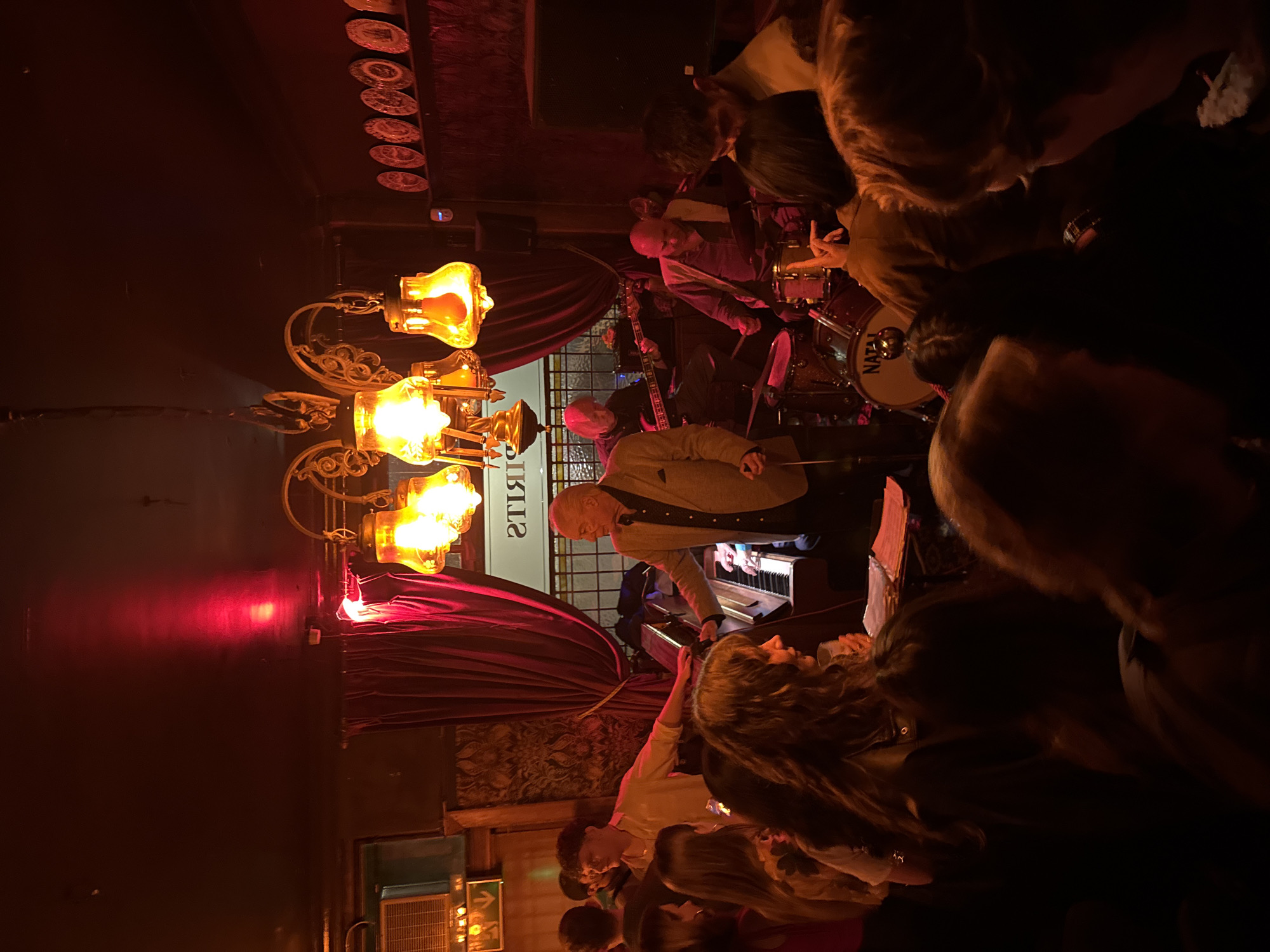 A dimly lit scene captures a live jazz performance in what appears to be a London bar. A band plays on a small stage, consisting of a drummer (whose drum reads "NATAL"), a bass guitarist, and a pianist in a light-colored jacket. The room is crowded with patrons, their faces obscured by the low light and the angle of the shot. An ornate chandelier with glass lamps hangs from the ceiling, providing one of the primary light sources. Maroon curtains frame the stage area. Plates adorn the upper wall, and a sign in reverse reads "SPIRITS." There are other light sources visible in the lower left corner. Patrons are densely packed in the foreground and on the right side, facing the stage.