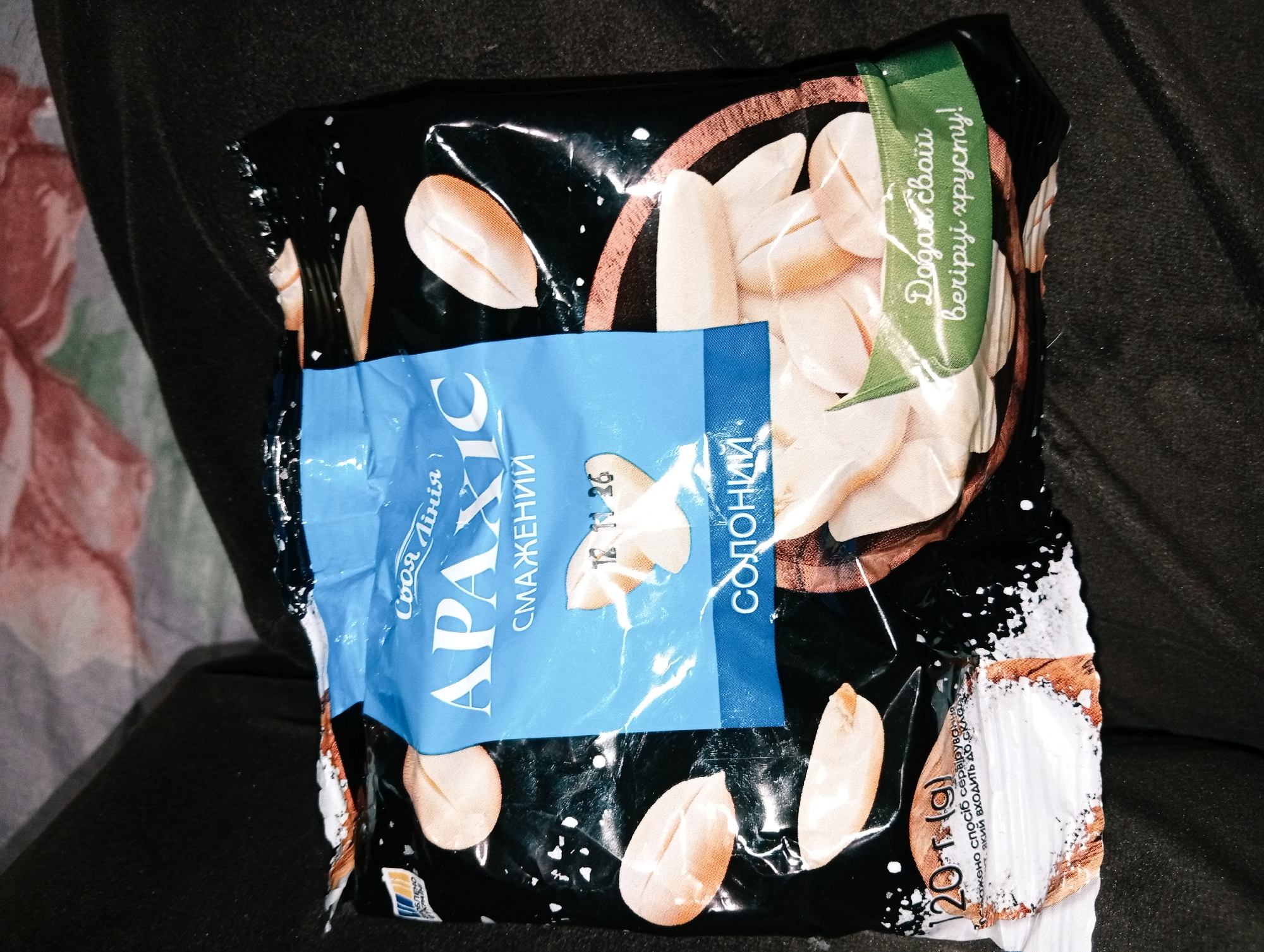 The image shows a partially opened bag of "Svoya Liniya" brand salted roasted peanuts ("Araxis" in Ukrainian) on a dark, possibly fabric surface. The bag is black with images of peanuts and a blue label. The date "12.11.26" is printed on the label. A green tag states "Add your own flavor!". The packaging indicates a weight of 120 grams. White salt is visible around the edge of the opened packaging. There is no indication of the image being taken in Poltava, Ukraine beyond the Ukrainian text on the package.