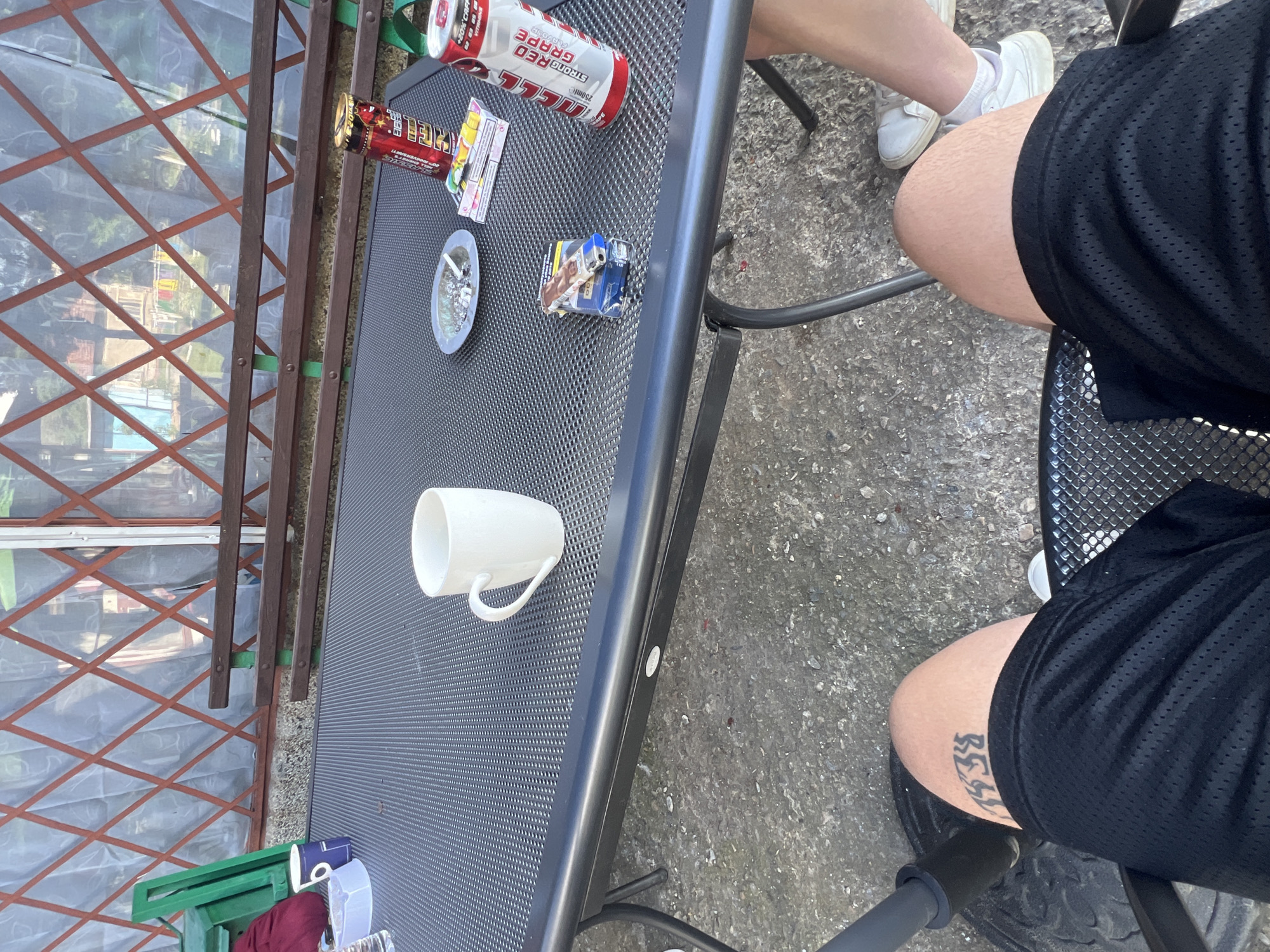 The image shows a table with various items on it, including a white mug, an ashtray with cigarette butts, a pack of cigarettes, and cans of a caffeinated beverage. A person wearing black shorts is seated at the table. The setting appears to be outdoors, possibly a patio or garden, based on the presence of a fence and the concrete ground. No specific indication of the location being Trifonovo, Bulgaria is visible.