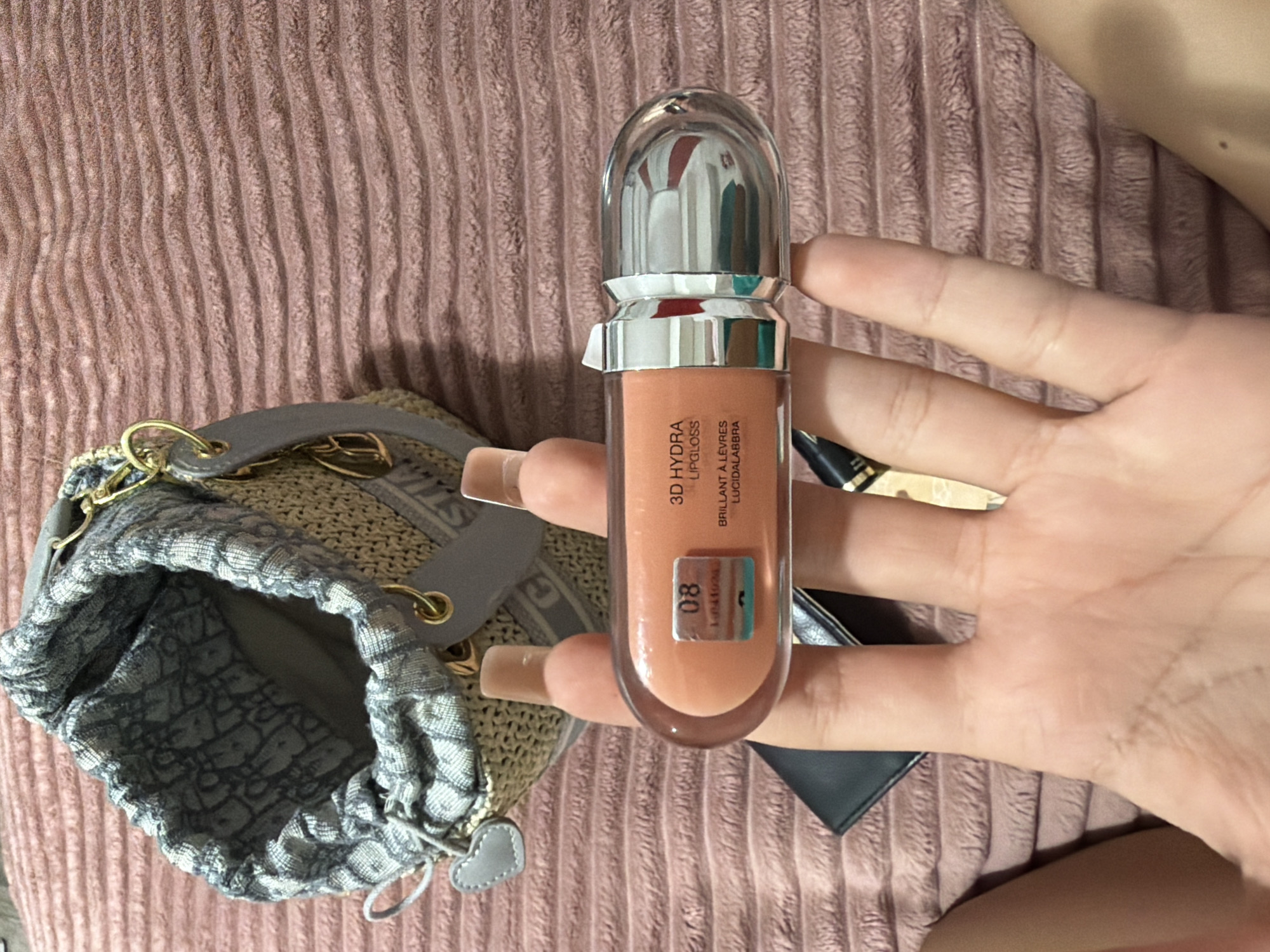 A close-up, high-angle shot captures a person's hand holding a Dior Lip Glow Oil in shade 08, "Pink Glow". The product is a translucent tube with a silver cap and contains a peachy-pink tinted oil. The background features a woven straw bucket bag with a grey Dior-patterned drawstring pouch inside, resting on a ribbed pink textile surface. The lighting is soft, creating a warm and inviting ambiance. There is no discernible connection to Digah, Azerbaijan in this image.
