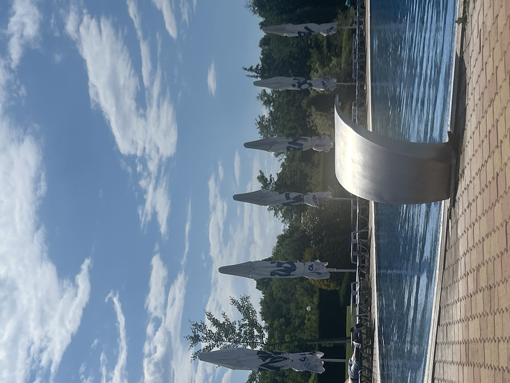 A person is lying on a lounge chair next to a swimming pool. The pool is a bright blue color and surrounded by a tiled walkway. In the background, there are several closed umbrellas with blue logos, lounge chairs, and lush green trees. The sky is a clear blue with scattered white clouds. A metallic waterfall feature is partially visible in the foreground.