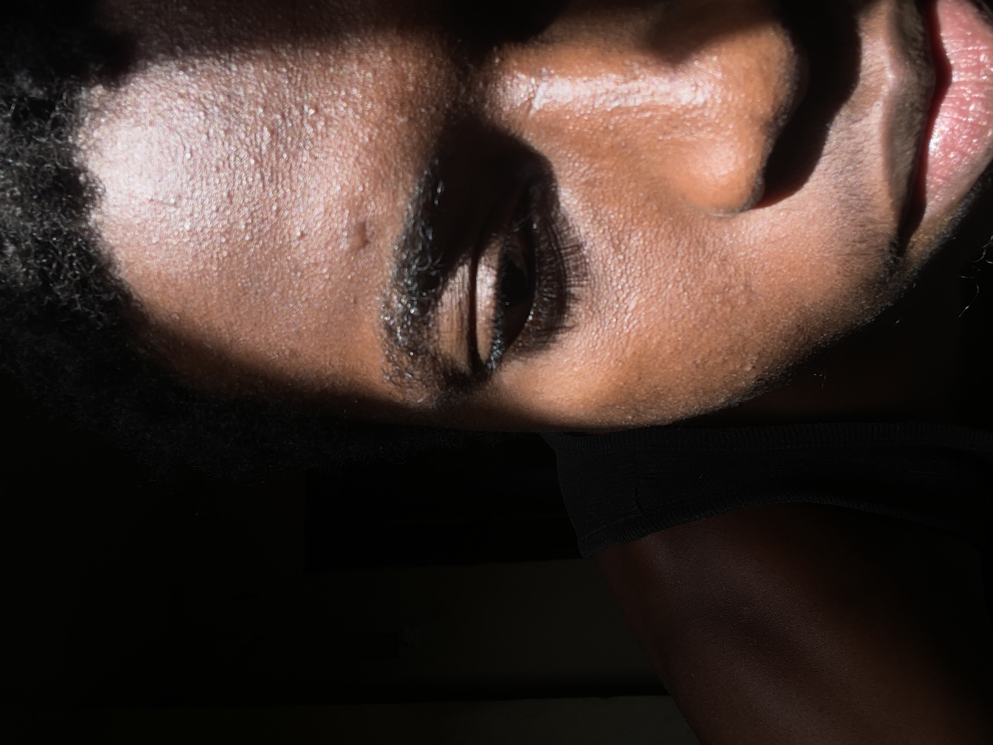 A close-up, slightly angled, and heavily shadowed portrait focuses on a dark-skinned person's face. The person's face is illuminated by a strong light source from above, creating stark contrasts of light and shadow. The person's eye is nearly closed and their lips are partially open. The person has dark hair visible at the top and left edge of the frame. A dark piece of fabric is partially visible below the chin, suggesting clothing.
There is no information available to determine the location to be Onitsha, Nigeria or any other location.