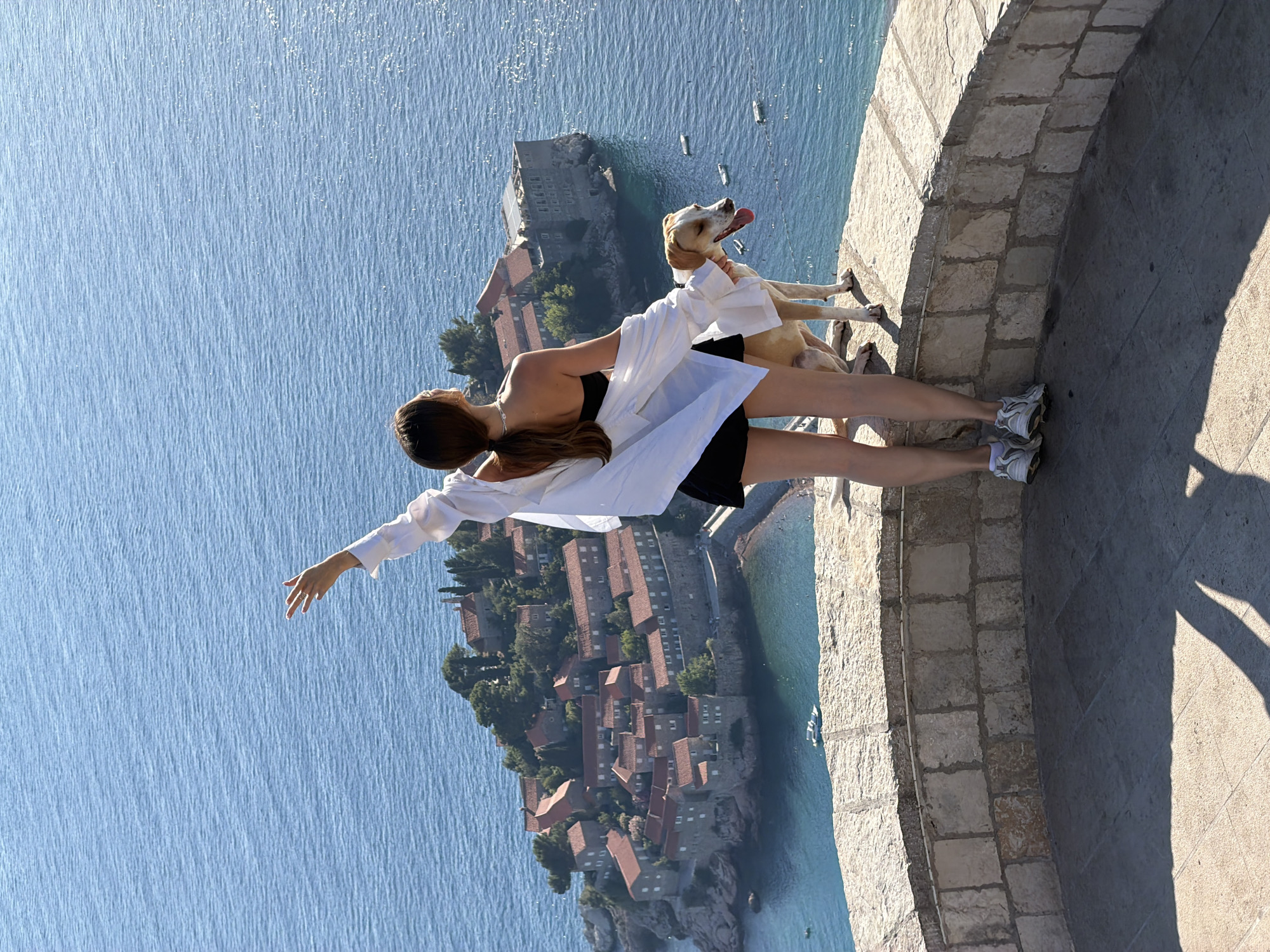 A woman and her dog stand on a stone balcony overlooking the island of Sveti Stefan in Montenegro. The woman, seen from behind, wears a black strapless top, black shorts, and a white open shirt. She has her left arm raised and her hand extended, as if gesturing. Her hair is tied in a ponytail. The dog, a medium-sized light brown and white breed, stands beside her, looking to its right with its tongue out. The island of Sveti Stefan, a fortified islet connected by a causeway, is visible in the background with its terracotta-roofed buildings and lush greenery. The sea surrounding the island is a deep blue. The balcony is constructed of stone blocks and has a curved wall. The ground in the foreground is paved, and shadows are cast across it. Several small boats are scattered in the water near the island.