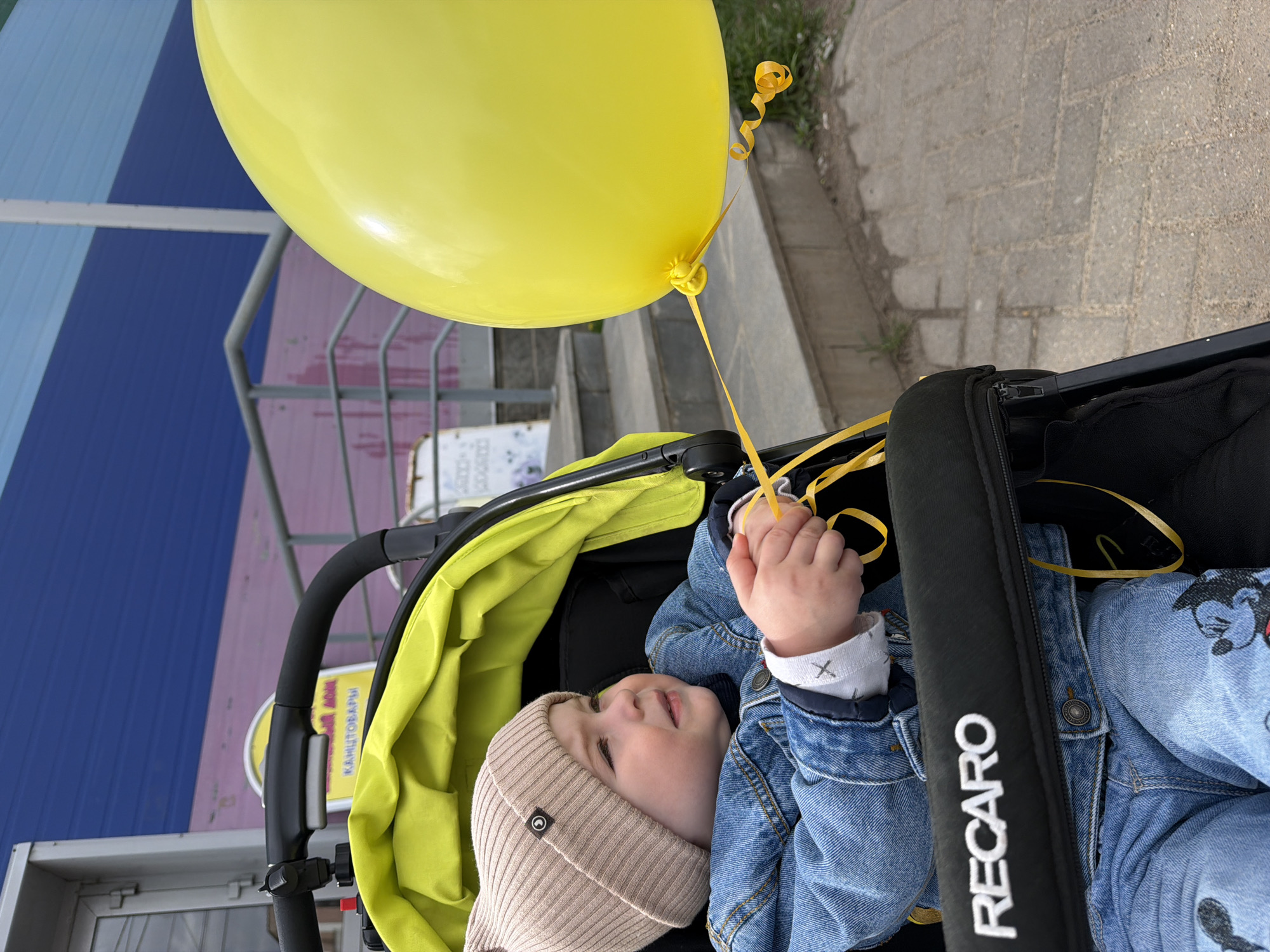 A young child, wearing a beige knit cap, a denim jacket, and denim pants with cartoon characters printed on them, sits in a stroller with a lime green interior. The stroller has a black frame, with "RECARO" stitched in white on the safety bar. The child is holding the string of a large yellow balloon. The background features a building with horizontal stripes of blue, pink, and grey, suggesting an urban setting. The presence of a sign with Cyrillic text indicates the possible location being Votkinsk, Russia. The ground is paved, indicating an outdoor scene.