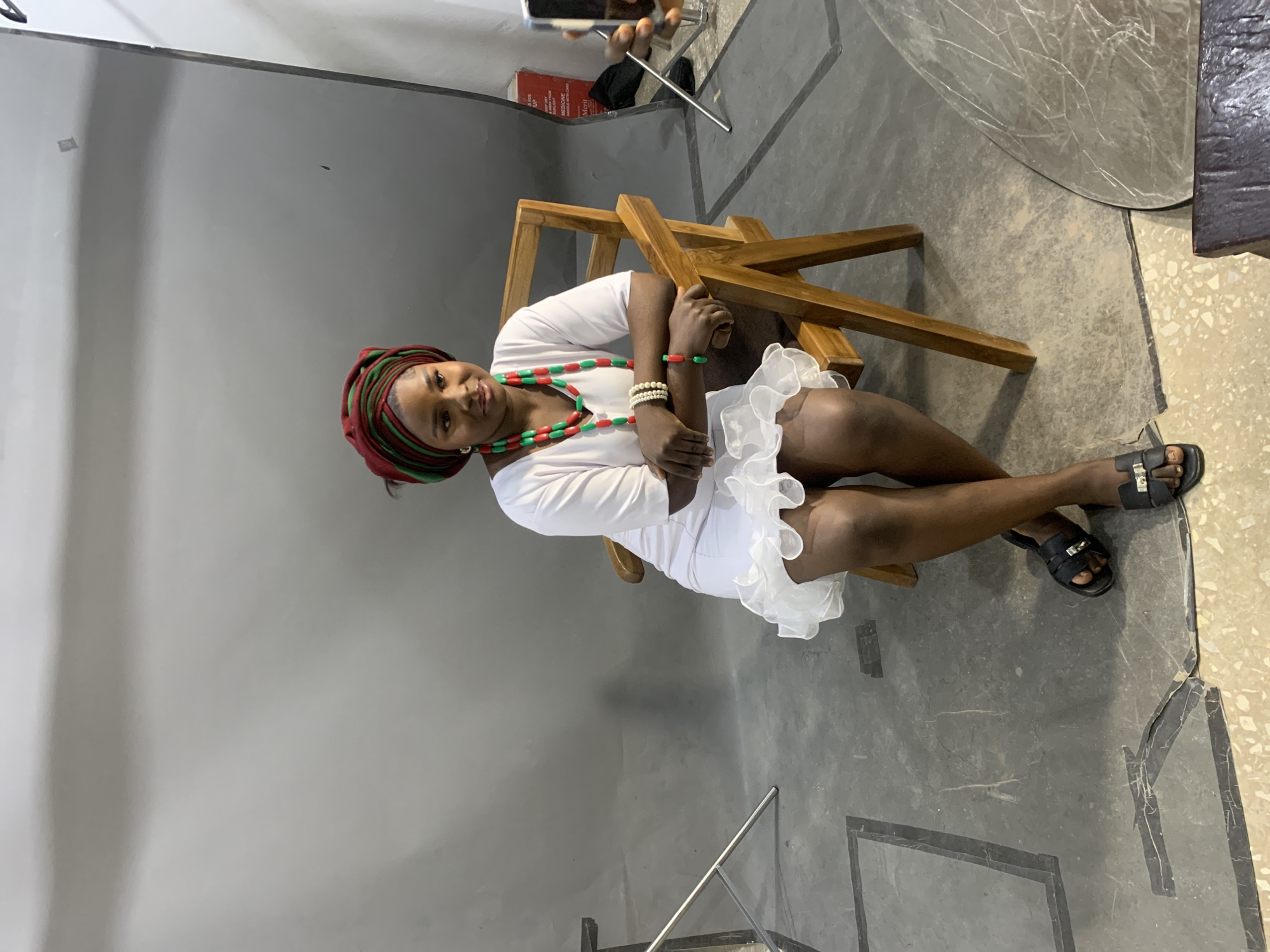 A young woman is seated on a wooden chair in a studio setting. She is wearing a white dress with a ruffled hem and a headwrap in shades of red and green. She has on beaded necklaces in similar colors and a bracelet. Her dark sandals have gold buckles. The background is a plain gray wall, and the floor is a mix of concrete and a lighter, speckled material. A large, circular reflector is visible to the right, suggesting a professional photo shoot. A hand holding a phone can be seen at the top of the image, possibly capturing the scene. The lighting appears to be studio lighting. The woman's expression is a slight smile, conveying a calm and poised demeanor. The overall impression is that of a posed portrait session. There is a partially visible red box with some text on it to the left of the woman.