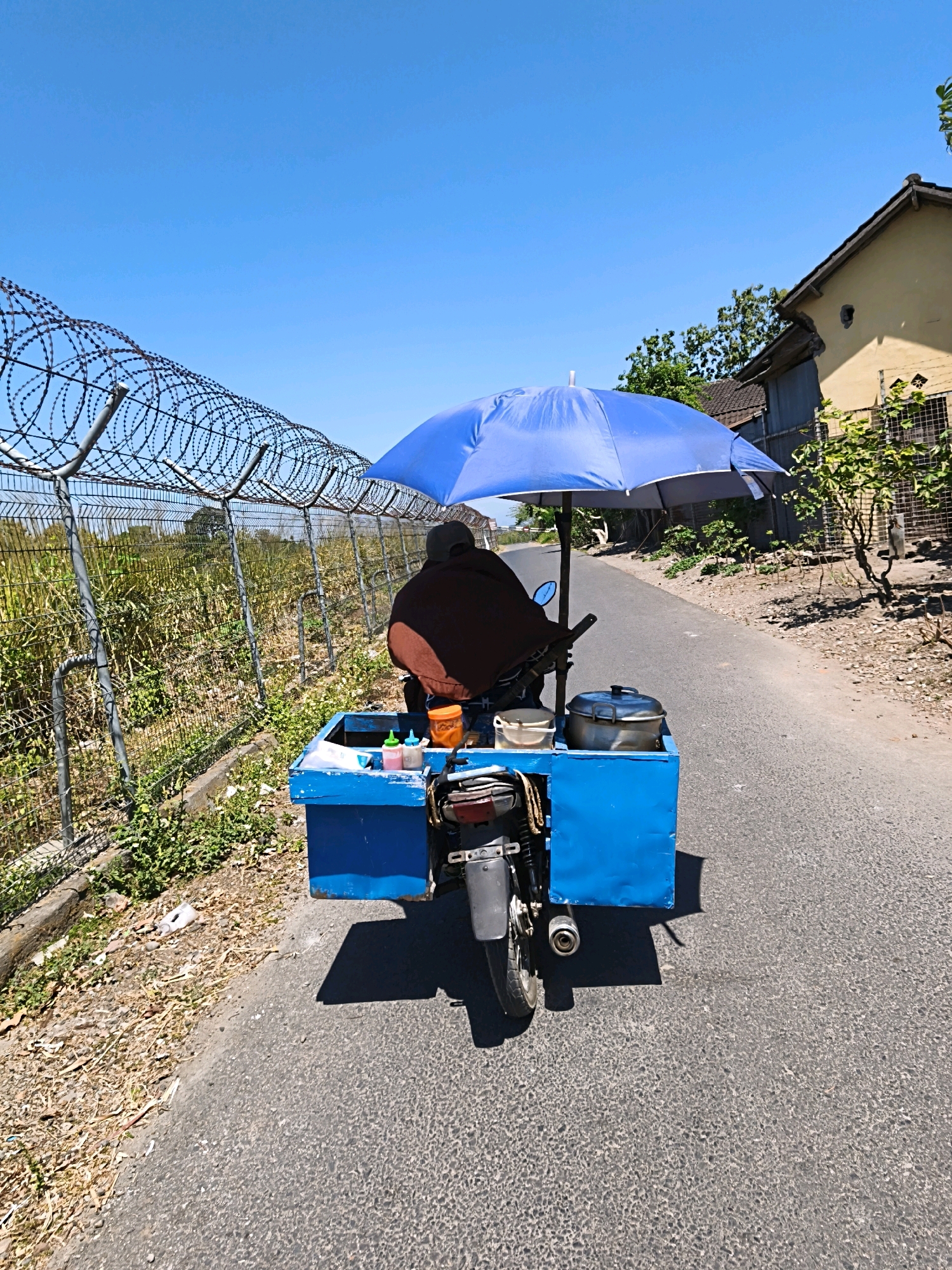 In Desa Dibal, Indonesia, on a paved road under a clear blue sky, a person wearing a brown garment and a dark cap rides a motorcycle modified with a blue cart. The cart is equipped with a large blue umbrella providing shade, and it contains several containers, including a large metal pot with a lid. Attached to the back of the cart are two small bottles with colored liquids. To the left of the road, a wire fence with barbed wire runs along the boundary of a field of dry vegetation. To the right, a yellow building with a dark roof and a wooden fence are visible. The scene captures a vendor on the move, navigating a rural road on a sunny day.