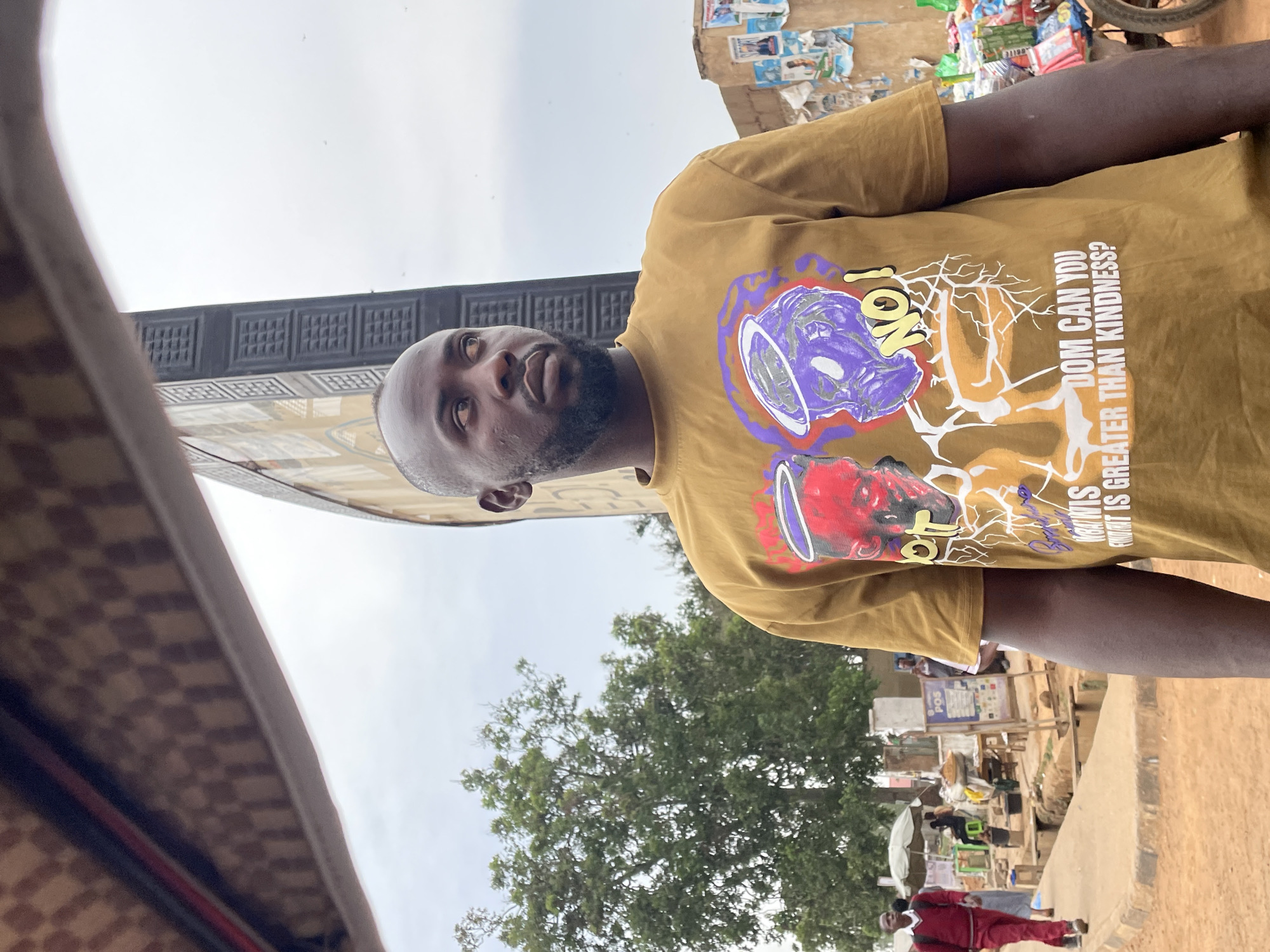 A medium shot of a bald-headed man, with a dark beard and mustache, is seen in Jos, Nigeria. He is wearing a tan shirt with a graphic design in purple, red and white. The shirt reads: "DOM CAN YOU" "WHAT IS GREATER THAN KINDNESS?" "1ON". He is looking up and to the left. The background includes a building with a brown and black square pattern, a green tree, and a street with a vendor's stall. A man wearing a red outfit is walking along the street. The sky is overcast.