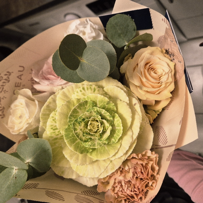 A bouquet features a variety of flowers. The focal point is a large ornamental cabbage with layers of pale green and white leaves. A peach rose sits beside it, complemented by smaller roses in white and light pink. Green eucalyptus leaves add texture and roundness. A peach-colored carnation peeks out at the bottom. The flowers are wrapped in patterned brown paper with "BEAU" visible.