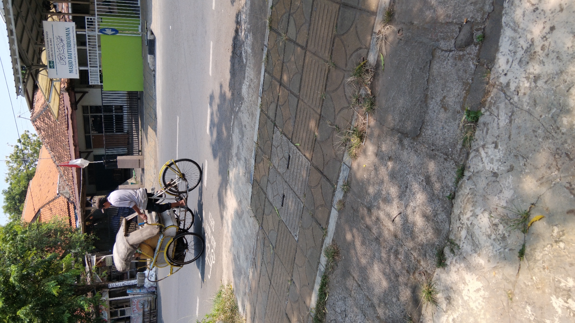 A man is riding a yellow pedicab on a street in Proyonangg, Indonesia. The pedicab is carrying a covered load. The man is wearing a white shirt and black pants. The street is paved with asphalt and has white lane markings. A sidewalk made of concrete tiles is visible on the right side of the street. In the background, there is a building with a red tile roof, a green wall, and a sign that reads "MASJID BAITURROHMAN". Trees and a flag are also visible. The scene is illuminated by sunlight, casting shadows on the street and sidewalk.