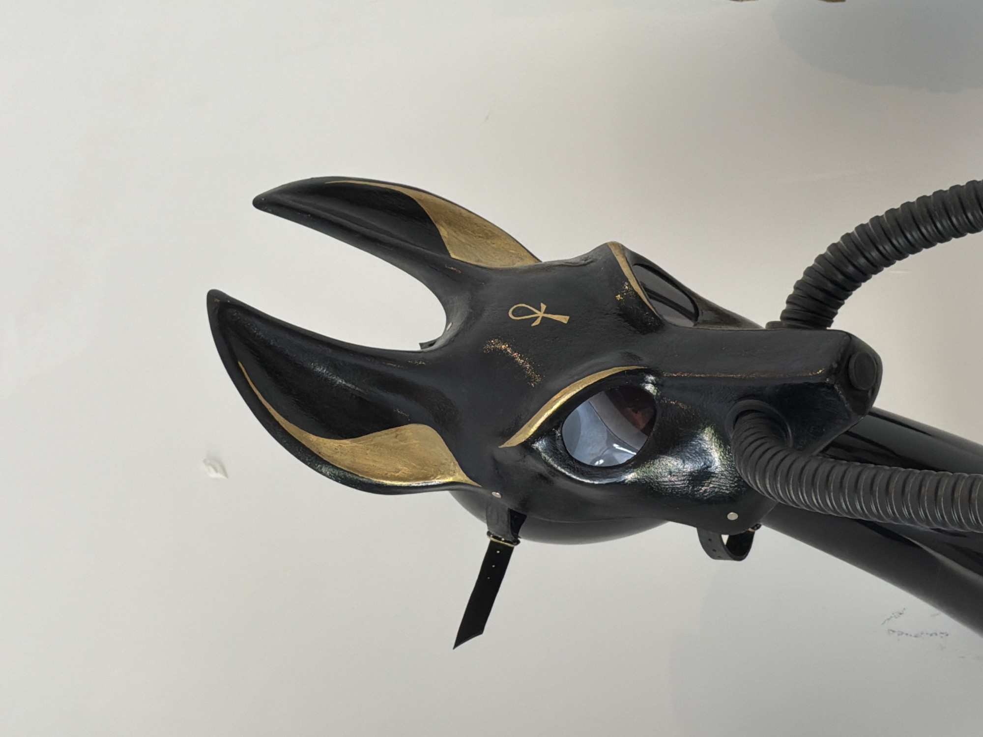 A black, Anubis-like mask with gold accents sits on a black stand against a white wall in Ko Samui District, Thailand. The mask features large, reflective eye lenses and an ankh symbol on its forehead. A black hose extends from the mask's snout, connecting to the stand. The wall has faint, dark markings.
