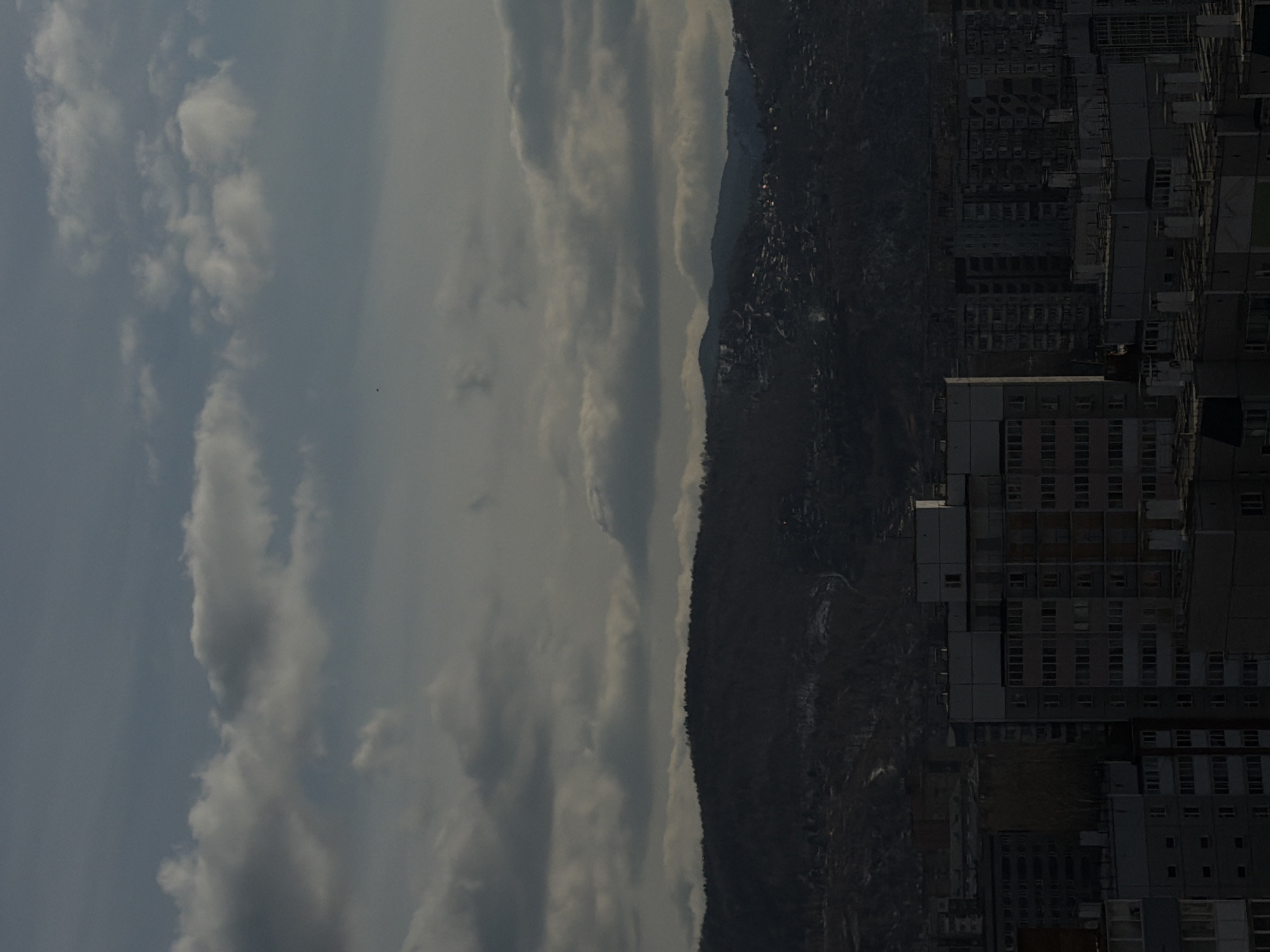 The image presents a panoramic view of a city nestled against a mountain range under an overcast sky, likely taken from an elevated vantage point in Krasnoyarsk, Russia. The perspective seems to be rotated, but when viewed correctly, the sky dominates the upper portion, a mountain range runs through the middle, and city buildings fill the lower and mid-ground.
The sky is heavily covered with thick, grey and white clouds, suggesting an overcast day or possibly a transition from morning to afternoon with diffused light. No direct sunlight is visible, contributing to a subdued and muted color palette across the scene.
A dark, imposing mountain range stretches horizontally across the mid-ground. The mountains appear densely forested with coniferous trees, giving them a dark green or brown hue. Hints of development or scattered lights can be discerned on the slopes, indicating human presence extending into the mountainous terrain.
In the foreground and mid-ground, the sprawling city unfolds. Numerous multi-story residential buildings, characterized by their functional, block-like architecture, dominate the urban landscape. These structures are primarily grey, white, and muted tones, with many windows, reflecting a typical urban environment in post-Soviet countries. The city seems to climb the lower slopes leading up to the mountains.
There are no visible people or explicit activities taking place, and no discernible text. The overall atmosphere is calm and somewhat somber due to the cloudy weather and subdued lighting.
