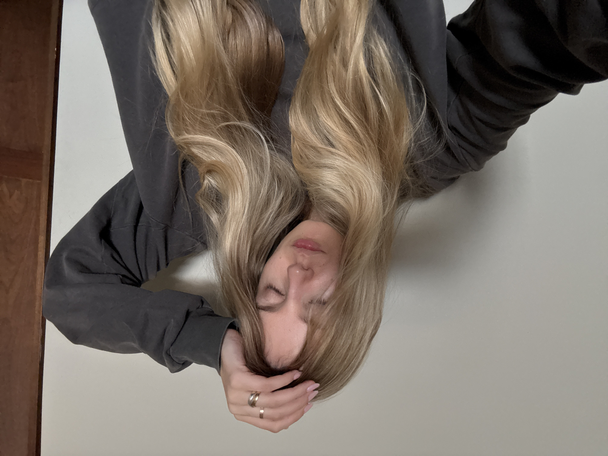 The image, taken in Mexico City, presents a close-up portrait of a person, likely a woman, in a state of rest or sleep. The individual has long, flowing, wavy blonde hair with subtle highlights, which frames their face. Their eyes are gently closed, and their lips are slightly parted, conveying a peaceful and serene expression.
They are dressed in a dark grey long-sleeved top, possibly a sweatshirt, with visible creases and folds in the fabric. One hand is positioned near their head, with fingers delicately resting near their temple. This hand prominently displays two rings: a double-banded or stacked ring on the ring finger and a simpler band on the middle finger. The fingernails are manicured with a light pink polish.
The background is simple, composed primarily of a plain, light-colored surface, likely a wall or floor. A narrow strip of a warm-toned wooden surface, possibly a door frame or piece of furniture, is visible on the left side of the frame. The overall atmosphere of the image is calm and tranquil. There is no visible text.
