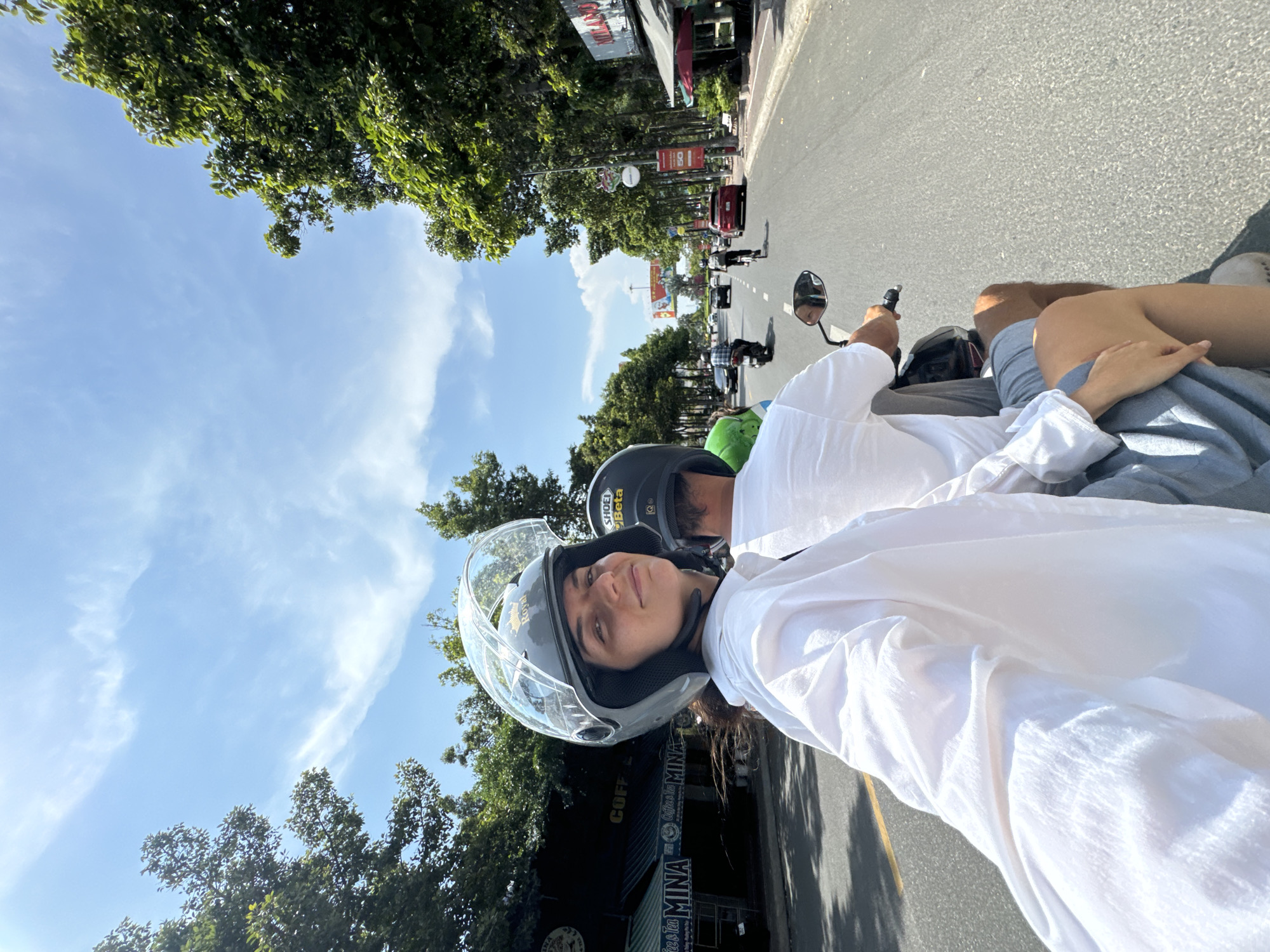 A low-angle, eye-level, medium shot captures a young woman and a man riding a motorcycle down a paved road lined with trees and buildings. The woman, positioned in the foreground and to the left, faces the camera with a slight smile, her head adorned with a grey helmet. She wears a loose-fitting white button-down shirt and light grey shorts. The man, seated behind her, is partially visible and wears a white t-shirt and grey shorts. He is operating the motorcycle, his hands on the handlebars. The motorcycle's mirror reflects a portion of the road ahead. The road is two lanes wide, with other vehicles, including cars, a bicycle, and another motorcycle, traveling in the same direction. Buildings, some with awnings and signage, are visible on the right side of the road, while lush green trees line both sides, creating a canopy overhead. The sky is a bright blue with scattered white clouds. A yellow line marks the edge of the road. The scene appears to be in an urban or suburban area, possibly Phu Thuy Ward, Vietnam, given the presence of tropical foliage and architectural styles.