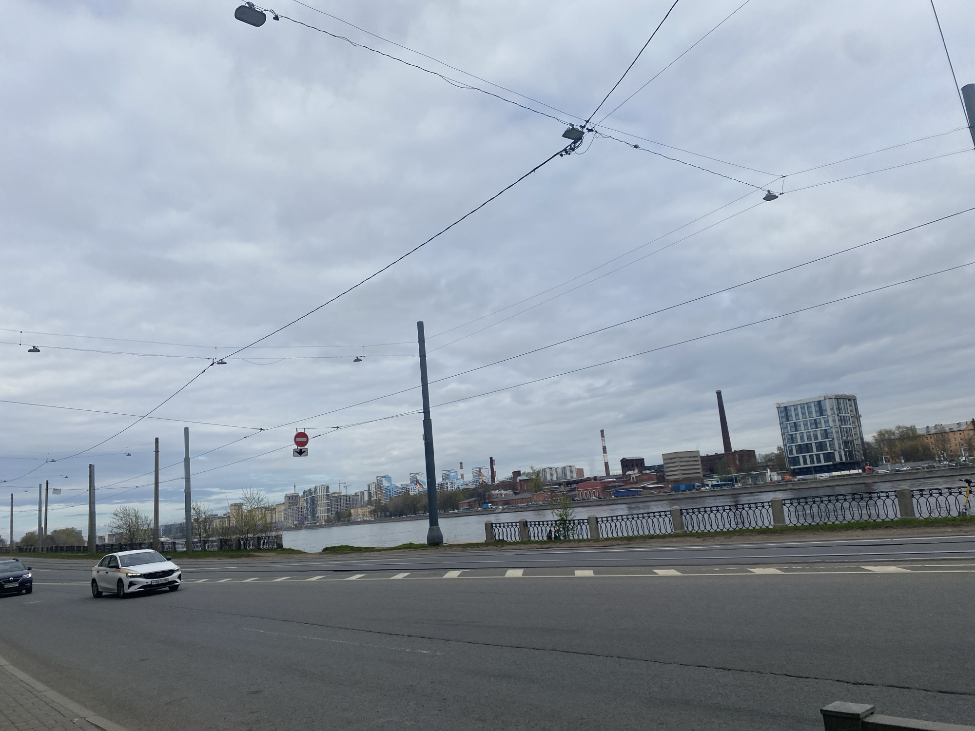 The image depicts a street scene in Saint Petersburg, Russia, likely along the embankment of the Neva River. The sky is overcast and gray. In the foreground is a multi-lane road with two cars: a white car driving to the right and a black car driving to the left. Utility poles and wires span the view above the road. Across the river, a cityscape is visible, including industrial buildings with chimneys, and modern residential or office buildings. A decorative iron fence runs along the riverbank. A stop sign is visible on a pole beside the river.