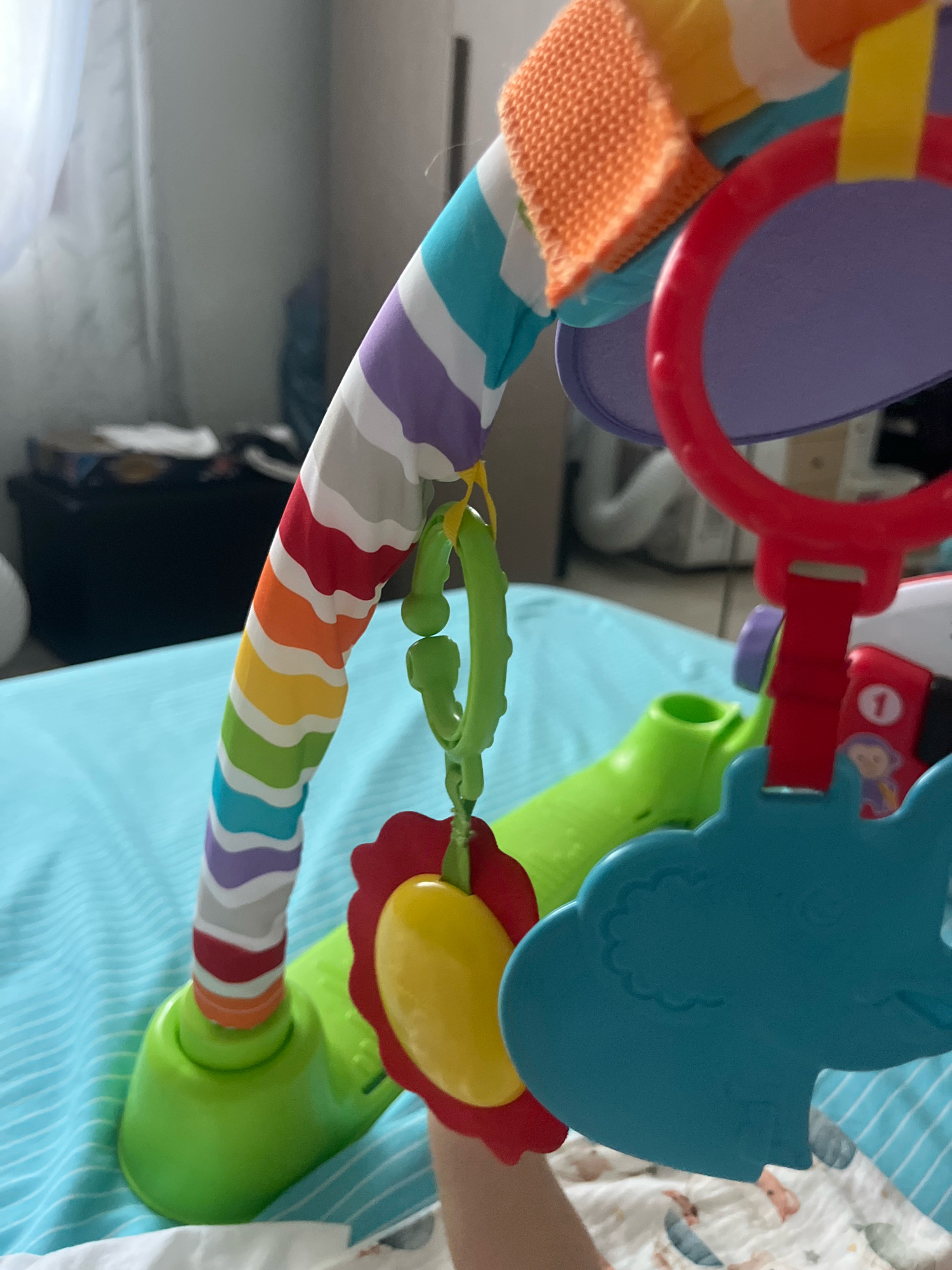 The image shows a close-up of a colorful baby gym. The gym has a rainbow-striped arch with various attached toys, including a green textured ring, a yellow and red flower-shaped toy, and a blue elephant. The gym is set up on a light blue, striped surface, possibly a play mat or bed. In the background, a black object, possibly a storage bin, is visible, along with sheer white curtains. The lighting suggests an indoor environment. There are no people clearly visible in the frame, though a small part of a baby's arm is visible near the bottom right corner. The scene does not provide any specific clues to its location within Zola Predosa, Italy.