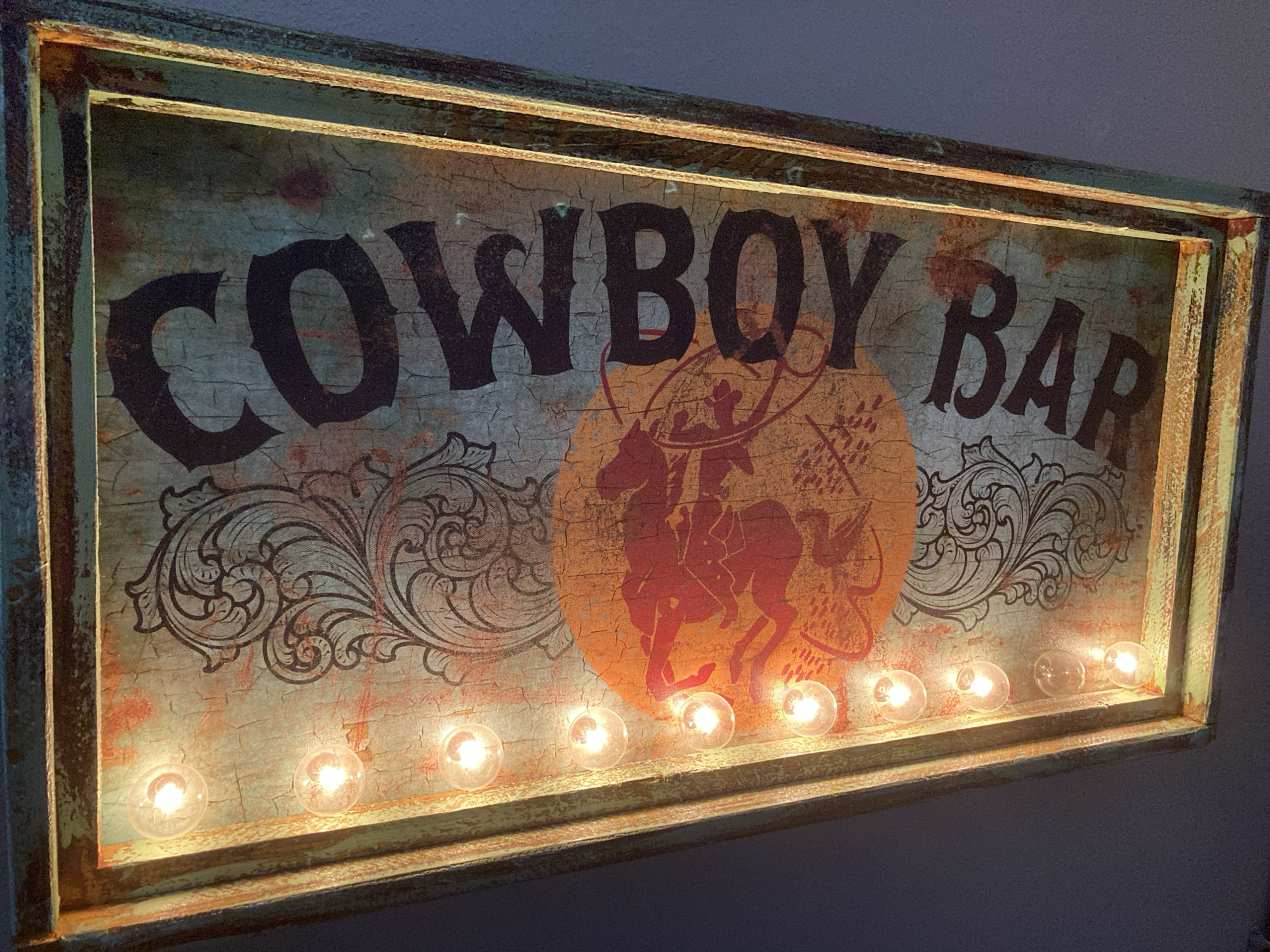 The image shows a close-up of a weathered, rectangular sign that reads "COWBOY BAR". The sign has a distressed, crackled texture with a teal and orange color scheme. A silhouette of a cowboy on a bucking horse, lassoing, is featured in the center, superimposed over an orange circular graphic. Ornate scrollwork is present on the left and right sides of the sign. A row of small, clear, illuminated light bulbs is mounted along the bottom edge of the sign, casting a warm glow. The sign is framed by a distressed, dark teal and orange frame. The background is a dark, textured wall. There are no people or animals depicted in the scene. This sign is likely a decorative item for a bar or home. The location in Round Rock, United States of America is not discernable from the image itself.