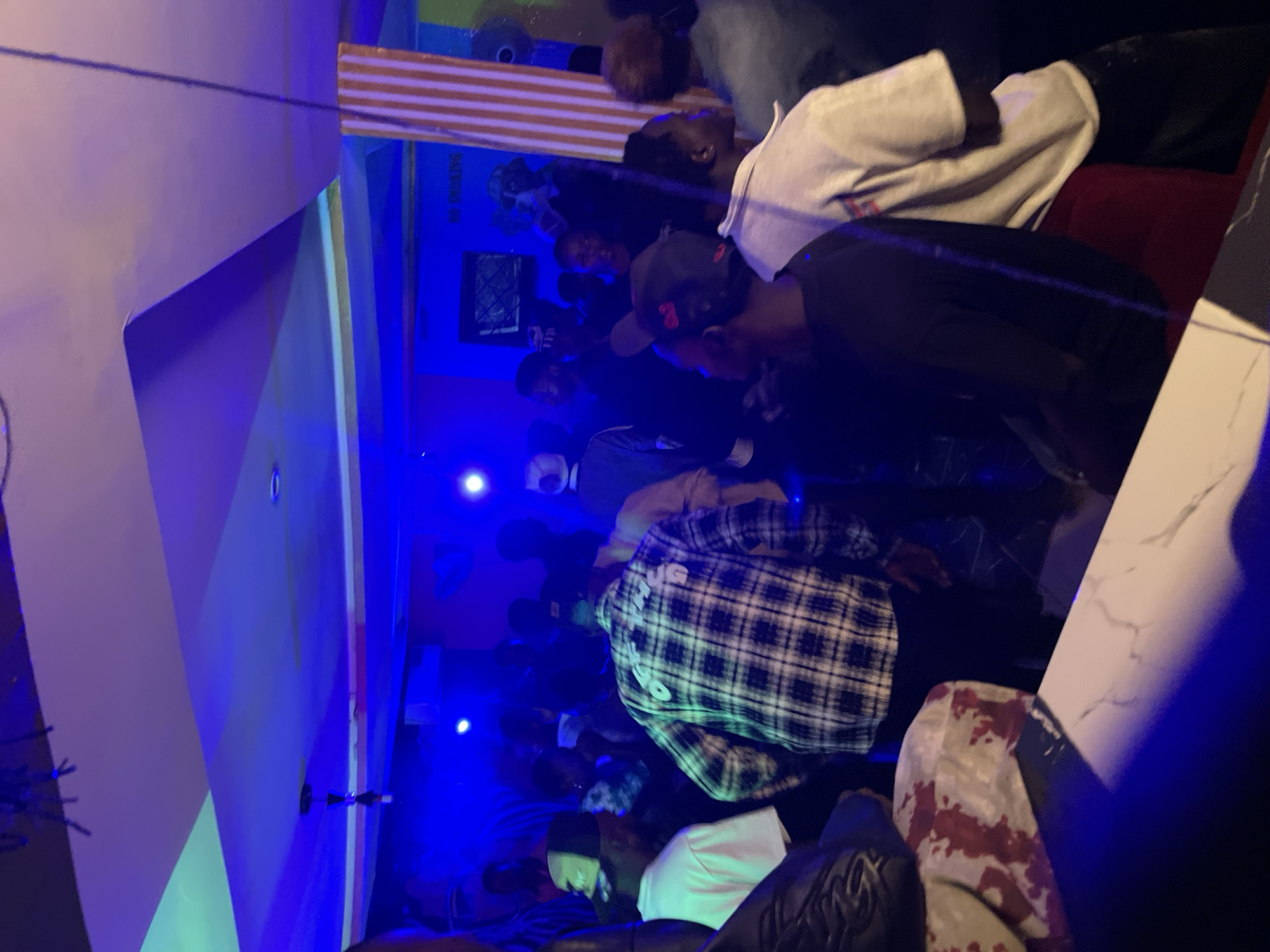 The image depicts an indoor gathering, likely a nightclub or a party, with patrons present. The environment is dimly lit, illuminated by a strong blue and purple ambient light, with some areas of green light visible on the ceiling. A prominent element is the ceiling structure, which features geometric angles and recessed lighting. In the foreground, a person is seen from the back, wearing a black and white plaid shirt. To their left, another individual in a white t-shirt and a cap is visible. Other individuals are scattered throughout the scene, some appearing to be dancing or socializing. A striped column, with orange and white stripes, stands to the right of the frame. A sign with the text "NO SMOKING" is partially visible on a wall in the background. The overall atmosphere suggests a lively, crowded social event. The location is identified as Durumi, Nigeria.