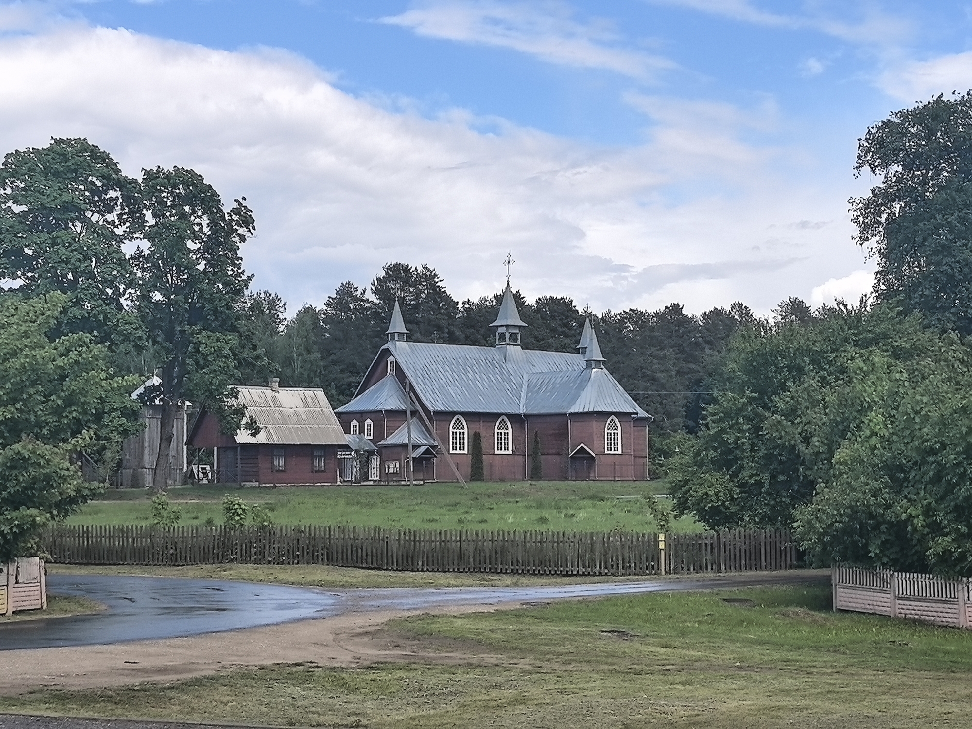 The image depicts a rural scene in Парэчча, Belarus, showcasing a red wooden church with a gray metal roof and three spires, the central one topped with a cross. The church is situated on a slightly elevated grassy field enclosed by a wooden fence. To the left of the church, a smaller red wooden building with a similar gray metal roof stands adjacent. The background is filled with dense trees. The foreground features a damp dirt road, with grass on either side and a wooden fence partially visible on the right. The sky is partly cloudy. No people are visible in the image.