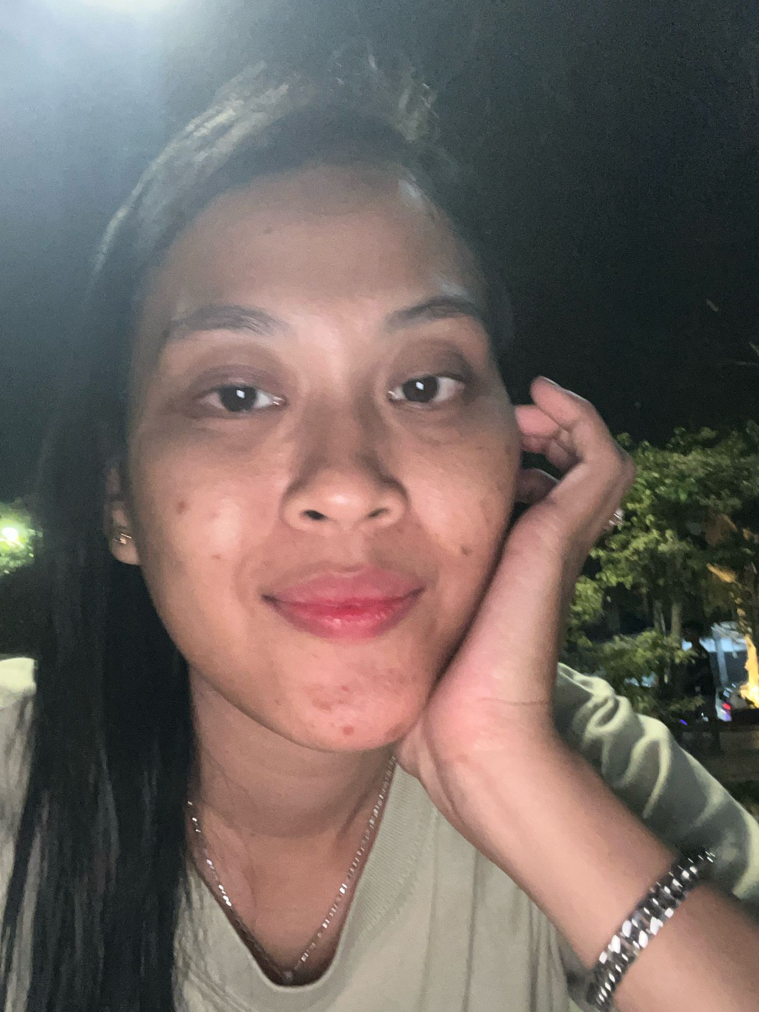 The image is a close-up, selfie-style photograph of a woman resting her head on her hand, taken outdoors at night in Kecamatan Pati, Indonesia. The woman has dark hair and is wearing a light-colored shirt and a silver necklace. She also has a silver bracelet on her wrist. In the background, there are trees and some blurry lights, suggesting an urban or suburban setting. The lighting is dim, with a bright light source visible in the upper left of the frame, possibly a streetlamp or building light.