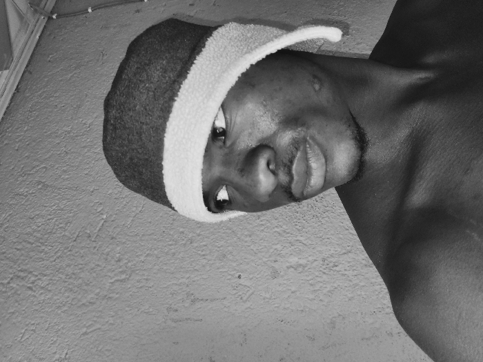 A black and white image depicts a male individual in Bukuru, Nigeria, captured from the chest up. The subject, with dark skin, wears a dark-colored beanie featuring a wide, light-colored, fluffy rim. He is bare-chested, revealing his shoulders and upper chest. He has a short mustache and a chin beard. His head is slightly tilted, and his eyes are directed upwards and to his left (viewer's left), with his lips slightly pursed. Visible marks are present on his left cheek/jawline and his right shoulder. The background is a textured, light-colored wall. In the upper right corner, a vertical structure, consistent with electrical conduit or wiring, is partially visible.