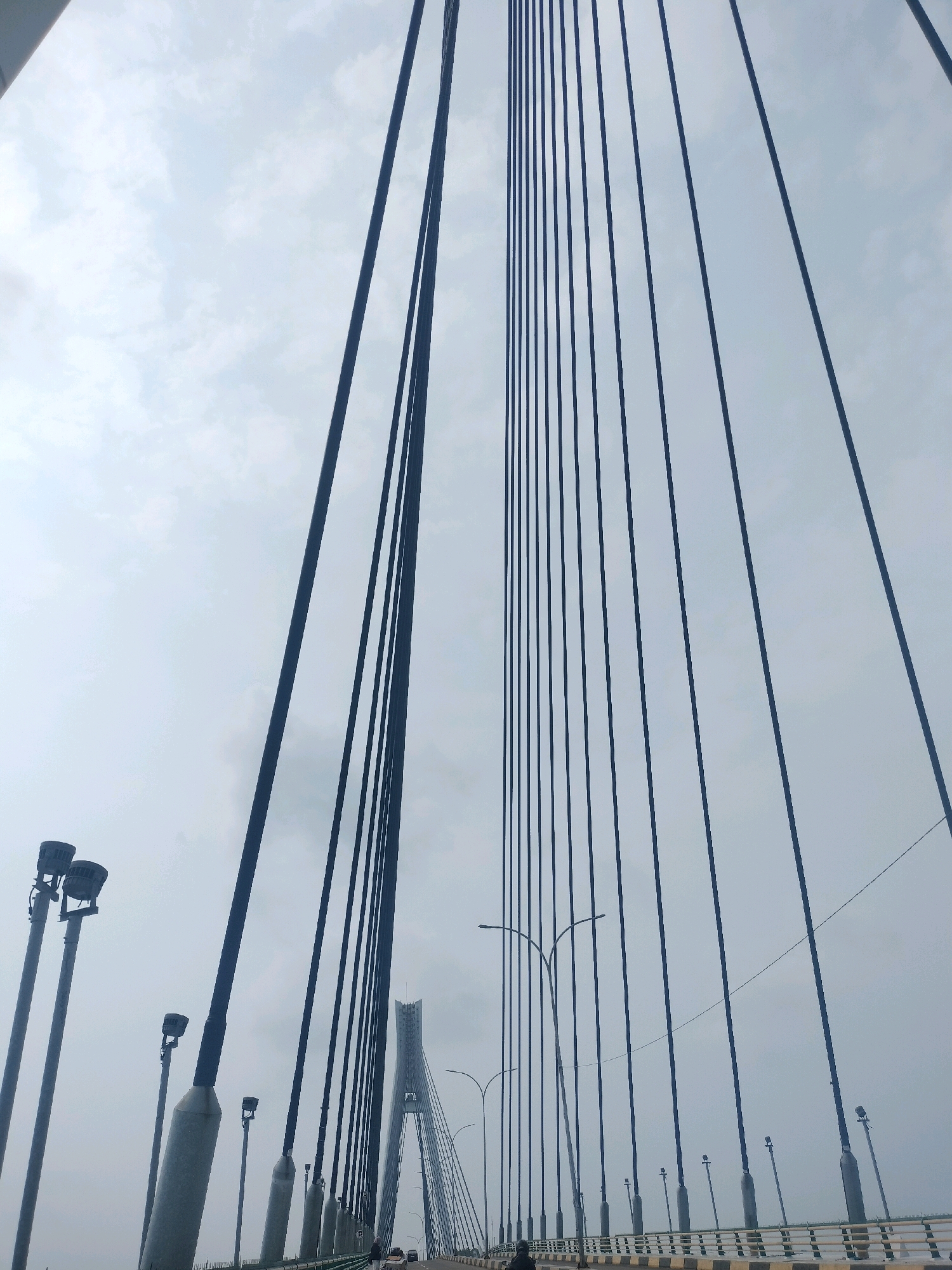 This is a ground-level, wide-angle photograph of the cable-stayed bridge known as the Barelang Bridge in Batam, Indonesia. The image is taken from the roadway of the bridge, looking upwards and slightly forward towards the bridge's suspension cables and pylons. The sky is overcast and a pale grey, contributing to the muted tones of the scene.
Dominating the frame are the thick, dark suspension cables that stretch diagonally upwards and outwards, creating a powerful geometric pattern against the sky. There are at least two sets of these cables visible, suggesting the perspective is from the midpoint or approach to one of the bridge's main towers.
On the left side of the image, several tall, slender lampposts are visible. They are grey and topped with clusters of lights. These lampposts appear to be spaced along the edge of the bridge's roadway.
Further in the distance, on the right side of the image, the distinctive silhouette of a tall, white pylon of the Barelang Bridge can be seen. This pylon is a landmark feature of the bridge's design, characterized by its A-frame or diamond shape at the top. Streetlights with curved arms are also present along the roadway in the distance, illuminating the path ahead.
The roadway itself is visible at the bottom of the frame, with a light-colored barrier and yellow markings. There are a few vehicles and at least two figures of people walking or standing on the roadway, indicating activity. The overall impression is one of scale and engineering, with the focus on the structural elements of the bridge against a vast, cloudy sky. The light is diffused, and there are no strong shadows. The scene suggests a typical day on the bridge, with traffic and pedestrians present.