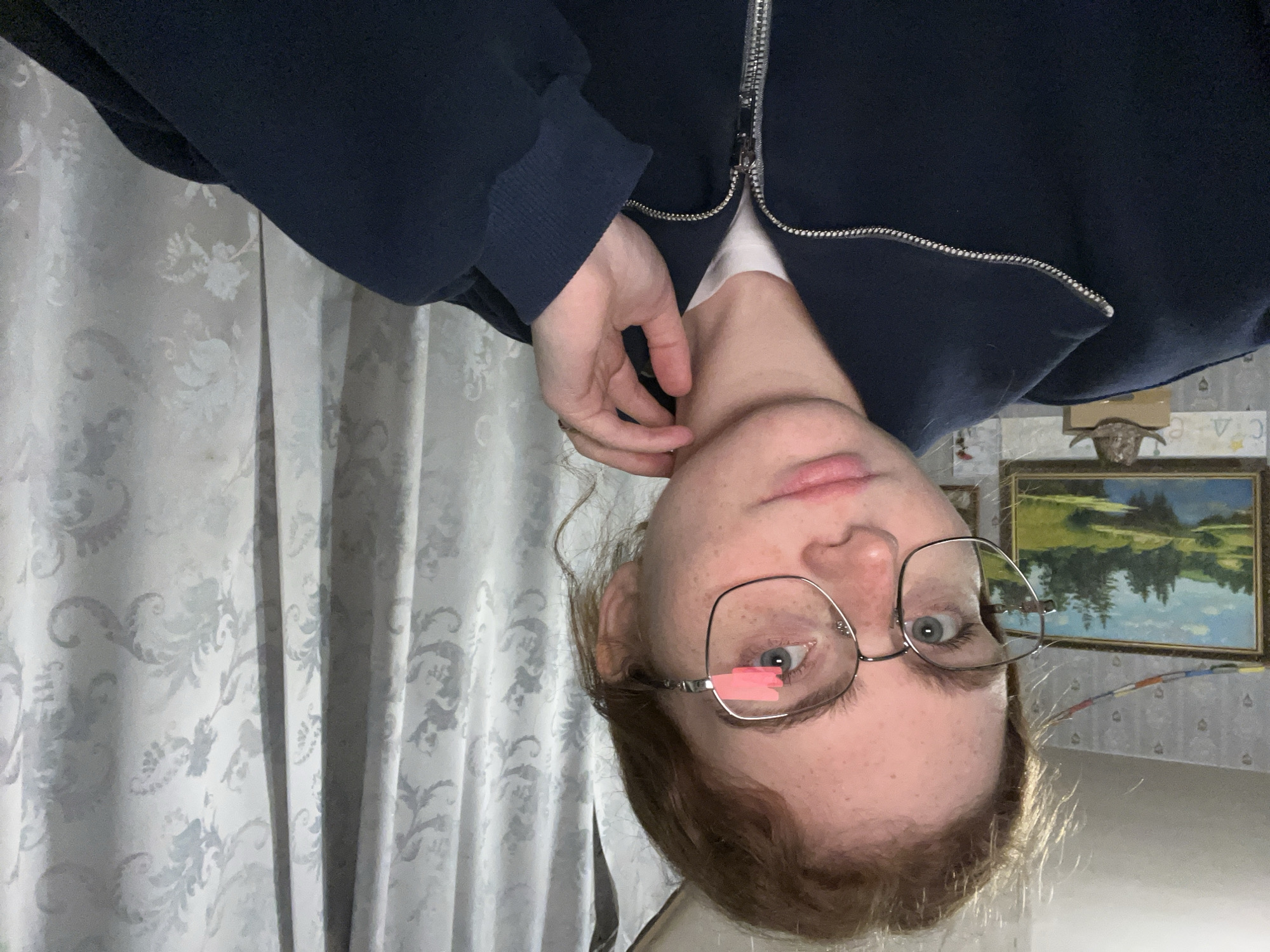 A young individual, likely female, with fair skin, light-colored hair, and blue eyes is featured in a medium close-up, looking directly at the camera. They wear square-framed metal glasses, with a small, bright pink rectangular object (possibly a sticky note or tape) affixed to the lower inner rim of the left lens. Their hair is loosely styled, with some strands around their face. They are dressed in a dark blue (navy) zip-up hoodie with a prominent silver zipper, worn over a white undershirt. One hand, adorned with a ring on a finger, is visible near their chin and collarbone area.
The background reveals an indoor setting. On the left, white curtains with a subtle floral or damask pattern are visible. On the right, the wall is covered with light-colored wallpaper featuring a repeating, subtle geometric pattern. A rectangular, framed landscape painting hangs on the wall, depicting green trees reflecting in blue water under a clear sky. A small, dark decorative object, possibly a bust or sculpture, rests on top of the painting's frame. The scene appears to be a room in a residence, potentially located in Новая Тура, Russia. The lighting is soft and ambient, characteristic of an indoor setting. The individual maintains a neutral and calm expression. No discernible text is visible in the image.
