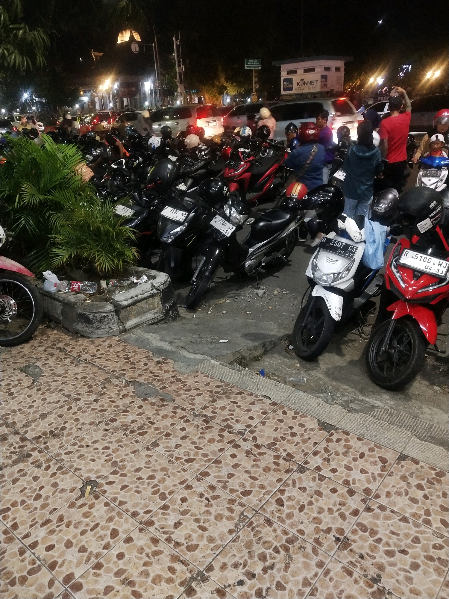 The image depicts a nighttime scene in front of a building with several motorcycles parked. The foreground is dominated by patterned tiles. To the left, a portion of a motorcycle wheel and fender is visible, along with a small, weathered concrete structure containing discarded plastic bottles and trash. Behind this structure, lush green plants are present. The middle ground shows a dense arrangement of parked motorcycles, with some individuals standing near them. One motorcycle, a white scooter with "Hello Kitty" stickers, has a license plate reading "R 2507 GS". Next to it, a red sport motorcycle has a license plate "R 518G WJ". Further back, more motorcycles and several cars are visible, illuminated by streetlights and building lights. A sign with "SIMPANG ALUN-ALUN" is visible in the background, indicating the location is near Alun-Alun, Purwokerto, Indonesia. Several individuals are standing around the motorcycles, some appearing to be talking or preparing to leave. The overall atmosphere is that of a busy area at night, likely a parking area or gathering spot.