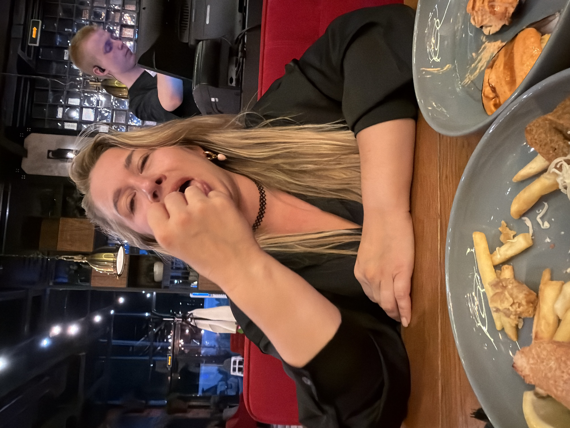 A medium shot captures a woman in a dimly lit restaurant, seated at a wooden table. She is leaning forward, with her right hand partially obscuring her face as she brings food to her mouth. Her eyes are closed, and her mouth is open in an expression of enjoyment. She is wearing a black v-neck top and a black choker necklace. Her blonde hair is loose, falling around her shoulders. In the foreground, two grey plates contain food, including french fries and what appears to be fried fish or chicken. In the background, a young man with short blond hair is visible, wearing earbuds and standing behind a counter with a laptop. The restaurant's interior features red seating and industrial-style lighting. The overall atmosphere is casual and social.