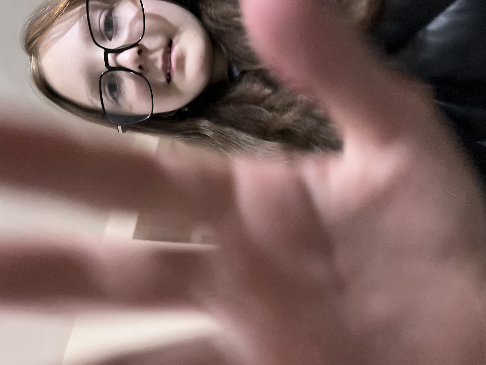 An angled, close-up photograph features a young person, likely a girl or young woman, with fair skin and light brown hair, positioned in the upper portion of the frame. She is wearing black-framed glasses and looks towards the camera with a neutral expression, her mouth slightly open.
The foreground is dominated by a large, out-of-focus, reddish-pink blur, which appears to be a hand or fingers positioned very close to the camera lens, partially obscuring the subject's face and creating a dynamic, abstract effect. Behind the subject, a dark, possibly black, garment is visible. The background is a plain, light-colored surface, indicating an indoor setting.
There are no discernible cues within the image to suggest the time of day or weather conditions. No visible text is present. The image does not contain specific visual elements to confirm the location in Chaikovsky, Russia.