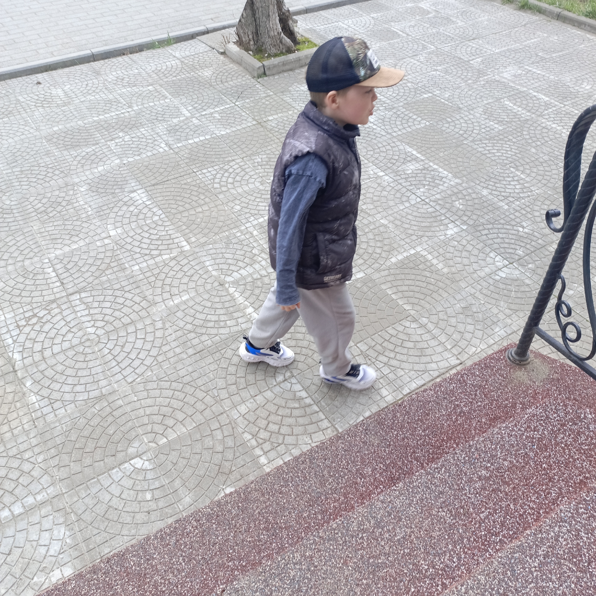 A child walks up outdoor steps in Unknown, Unknown. The child is wearing a baseball cap, a dark vest, a long-sleeved shirt, gray pants, and white sneakers with blue accents. The steps are made of a red, textured material, and there is a black wrought-iron railing on the right side of the image. The sidewalk leading to the steps is paved with gray tiles in a circular pattern. A tree is visible in the background.