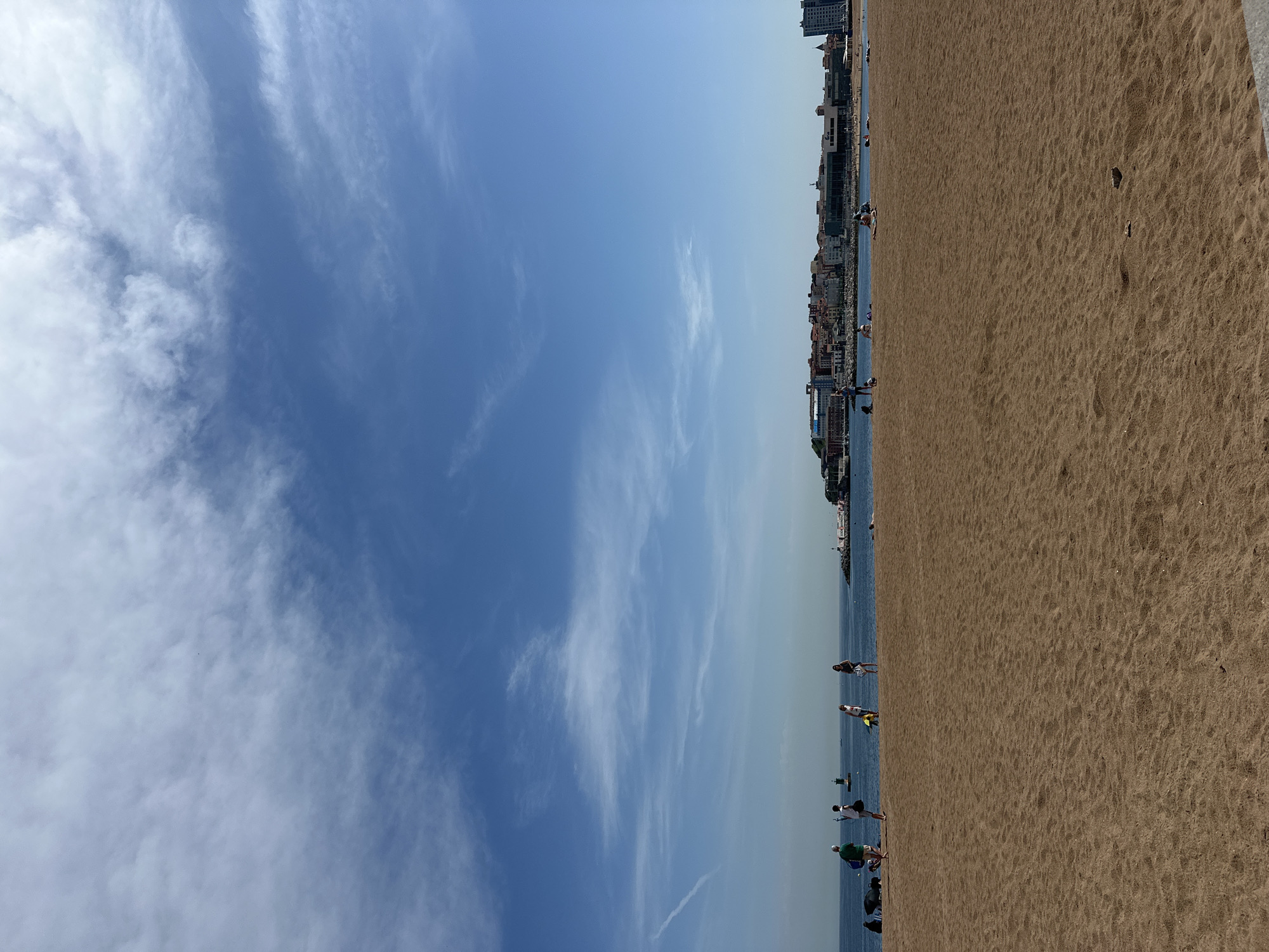 The image depicts a beach scene in Gijón/Xixón, Spain, characterized by a wide expanse of sand occupying the right side of the frame and a blue sky with wispy clouds dominating the left. A line of buildings is visible along the beach’s edge, delineating the city’s urban landscape. The horizon is positioned slightly above the center of the image.
People are scattered along the beach, engaging in various activities. Some are walking along the shoreline, while others appear to be standing or interacting in small groups near the water’s edge. The environment is brightly lit, suggesting a sunny day. The sea appears calm with a clear sky.