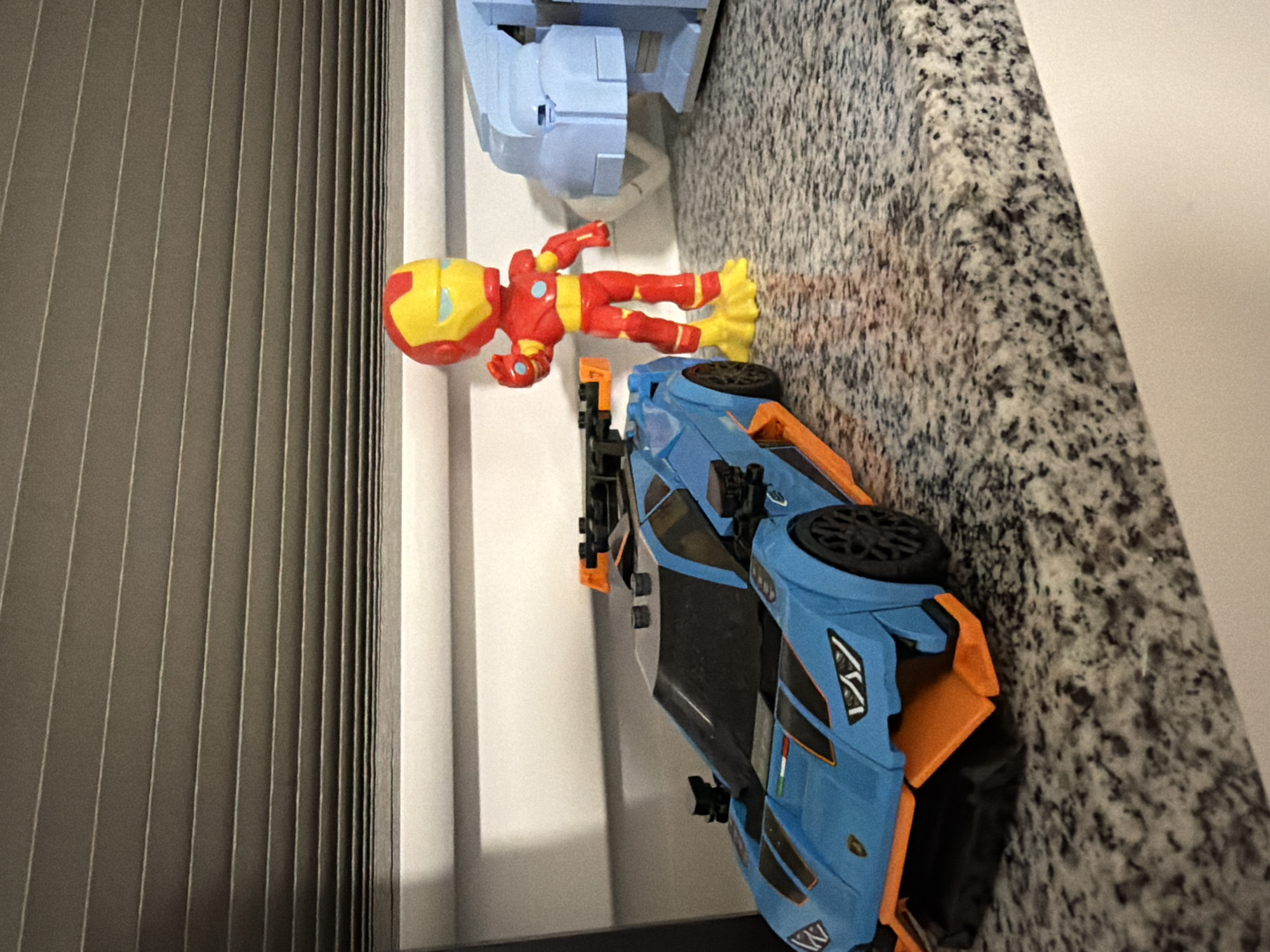 The image shows toys on a speckled countertop. A blue and orange toy car with a black spoiler is on the left. To the right of the car, a small red and yellow figure resembling Iron Man is standing, with its left hand raised. Behind Iron Man, a light blue toy building is partially visible. The countertop has a granite-like pattern. In the background, horizontal blinds are visible against a white wall. The image appears to be taken indoors, with no indication of the city of Sankt Augustin, Germany.